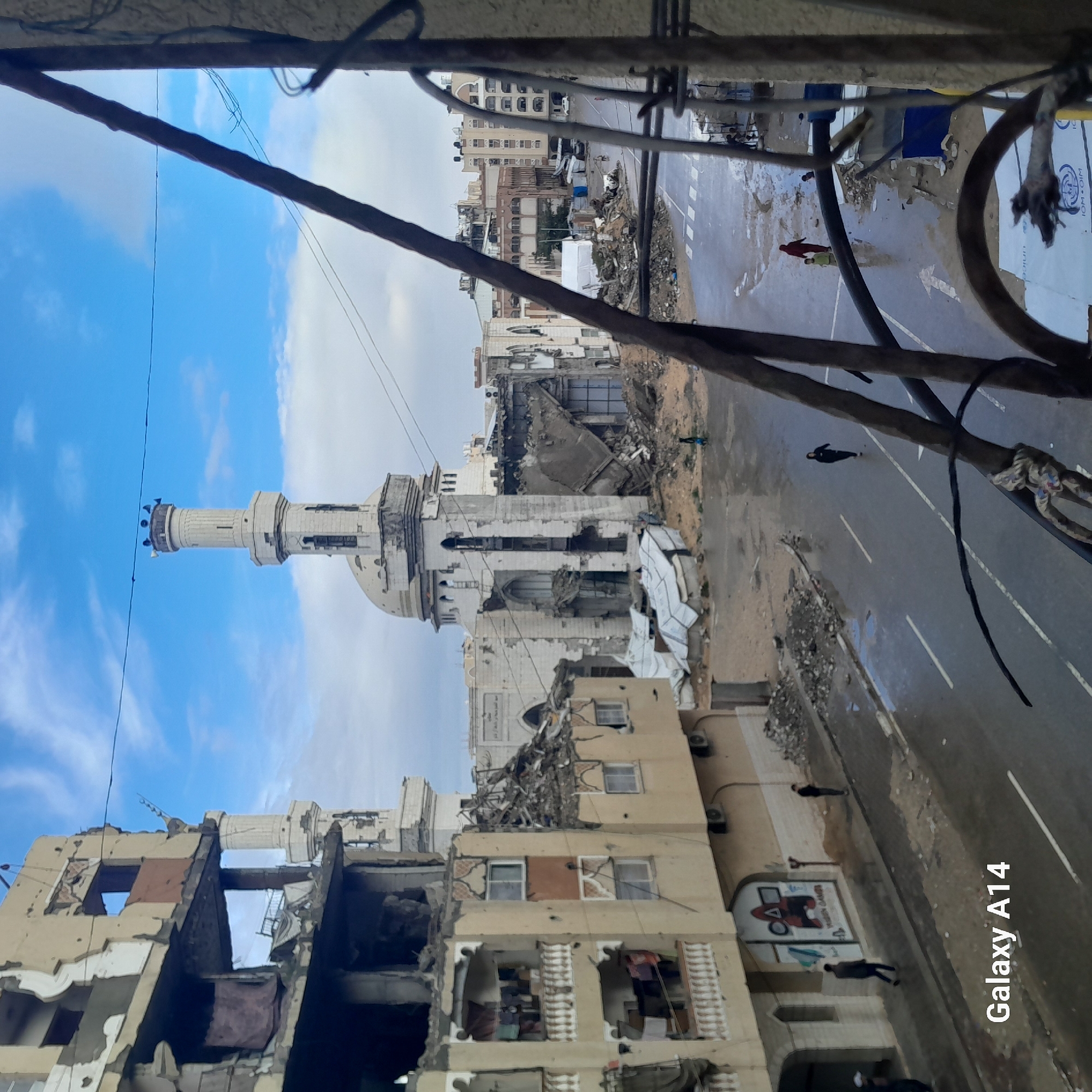 An elevated view captures a street scene in Khan Yunis, Palestinian Territory, under a partly cloudy sky. The central feature is a heavily damaged white mosque with a prominent minaret, still largely intact, fitted with loudspeakers. The mosque's dome and main structure exhibit significant collapse and structural compromise, with large sections of walls and roof missing.
To the left, multi-story buildings show extensive structural damage, including exposed interiors and collapsed balconies. Laundry hangs on several balconies, suggesting recent or ongoing habitation despite the damage. Graffiti, including a "Squid Game" motif, is visible on a lower section of one building.
The wet asphalt street below, marked with white lines and a pedestrian crossing, indicates recent precipitation. Piles of rubble and debris line the street and surround the damaged structures. Several individuals are present on the street: one person in dark clothing walks alone in the middle distance, another in red walks with a small child in yellow further down, and two individuals are on the left sidewalk near the damaged buildings.
In the background, further damaged buildings and what appear to be white temporary shelters or tents are visible. A white vehicle is partially discernible in the far distance. Metal bars, possibly from a window or balcony railing, are prominent in the foreground on the right, framing the scene. A partial view of a sign with "IOM OIM" is visible on the ground in the bottom right corner.