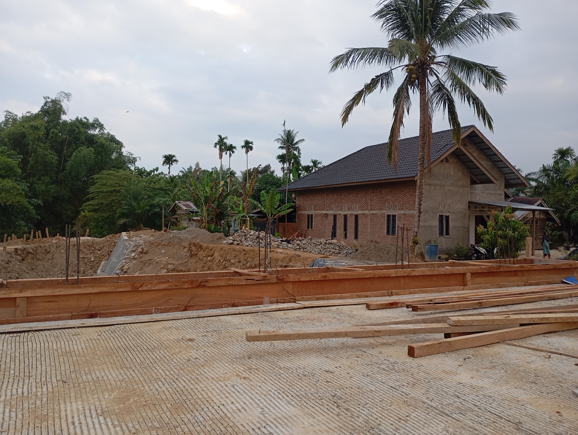 The image depicts a construction site in Blangraleu, Indonesia. In the foreground, a concrete foundation with parallel grooves is visible, overlaid with several wooden planks. A wooden formwork runs along the edge of the foundation, with exposed rebar extending upwards.
Behind the formwork, there is a large mound of dirt, likely excavated material, with some rocks and a concrete structure at the base. To the right of the dirt mound, a partially constructed brick building with a dark tiled roof stands prominently. This building has several windows and appears to be unfinished. A tall coconut palm tree with green fronds and a few coconuts grows next to the building.
The background reveals a lush, tropical environment with dense green vegetation, including bamboo and various other trees. Several smaller structures, possibly houses or sheds with corrugated roofs, are scattered among the trees. The sky is overcast with a muted light, suggesting it might be late afternoon or an early morning. No people are clearly identifiable in the immediate vicinity of the construction work. The overall scene conveys a sense of ongoing construction in a rural or semi-rural tropical setting.