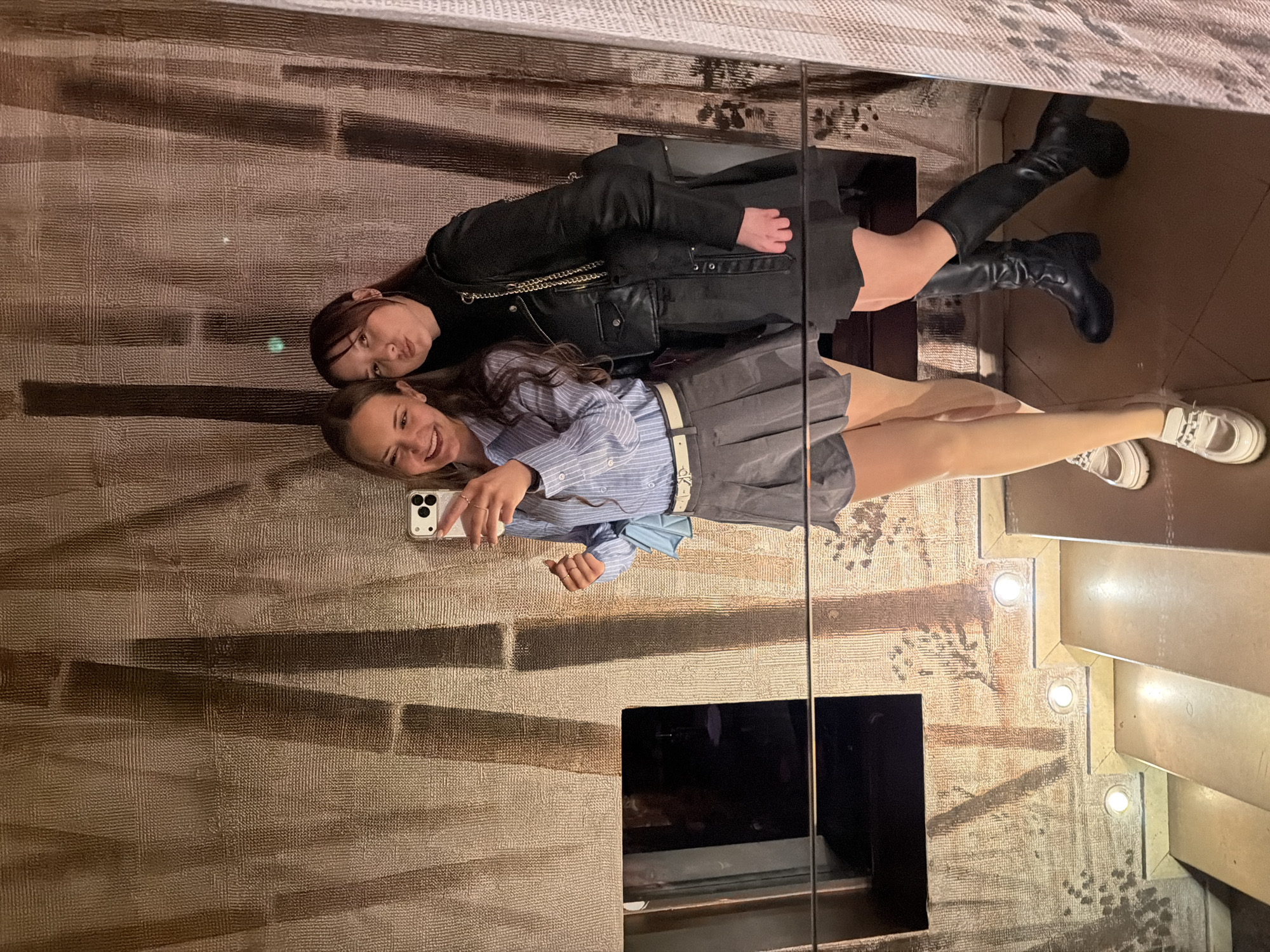 Two young women are posing for a selfie in front of a large mirror. The woman closer to the camera is holding a white smartphone. She is wearing a blue striped shirt, a gray pleated skirt with a white belt, and white sneakers. The other woman is behind her, wearing a black leather jacket, a black turtleneck, and black knee-high boots. The background consists of a textured wall with linear patterns and a mirrored surface reflecting parts of a staircase with recessed lighting. No specific location in Sofia, Bulgaria can be determined from the image.