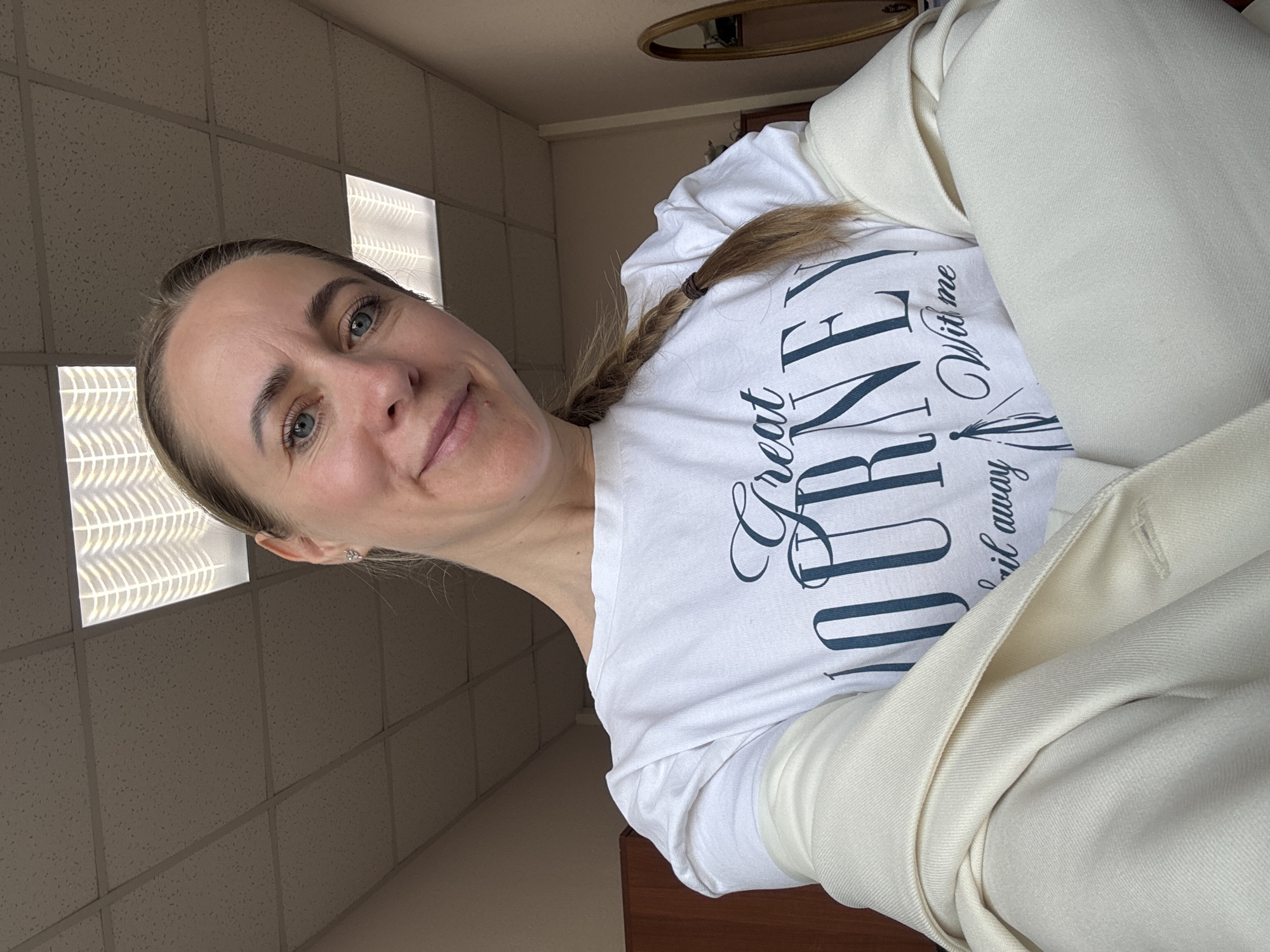 The image depicts a woman with fair skin, blue eyes, and blonde hair styled in a braid, posing for a selfie. She is wearing a white t-shirt with the phrase "Great Journey & Sail away With me" and a light-colored jacket. The background consists of a drop ceiling with fluorescent light panels, suggesting an office or institutional setting. Based on the prompt, this location is in Chaikovsky, Russia.
