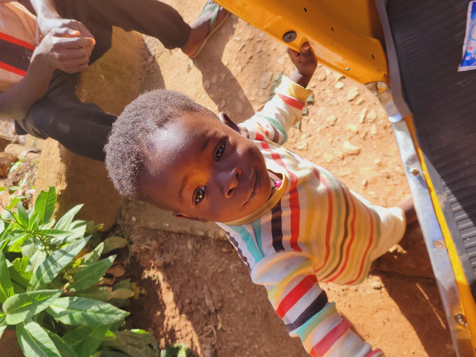 In Bukuru, Nigeria, a young child with dark curly hair and brown skin, wearing a striped long-sleeved shirt, looks towards the camera. The child's left hand rests on a yellow object, possibly part of a vehicle or structure. In the background, a person wearing dark pants and a striped t-shirt is seated, their hands clasped. The environment appears to be outdoors, with reddish-brown soil and green foliage visible. The lighting suggests it is daytime, with shadows cast on the ground.