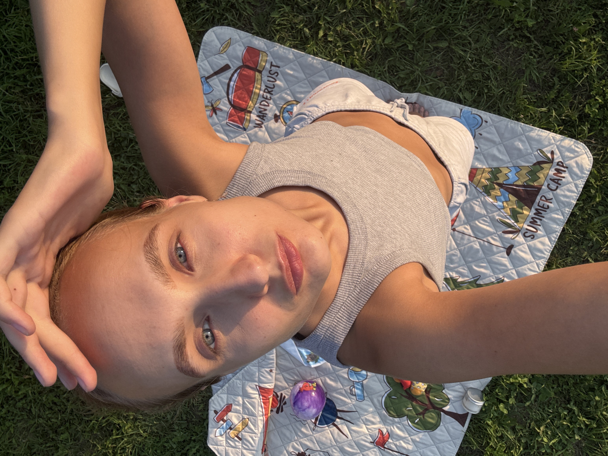 A high-angle, selfie-style shot captures a young woman lying on a picnic blanket on grass, illuminated by warm, low sunlight. The woman is seen from the chest up, with her head tilted back and her left hand resting on her forehead. She has light-colored hair, blue eyes, and fair skin with visible makeup on her lips and eyebrows. She is wearing a grey, ribbed tank top and white shorts. The picnic blanket has a quilted pattern with cartoonish drawings of camping-themed elements, including tents, trees, flags, and the text "WANDERLUST" and "SUMMER CAMP." A round, purple object, possibly a ball or toy, is visible on the blanket to the left of the woman. The background is green grass. The location is specified as the village of Podshivalovo, Russia.