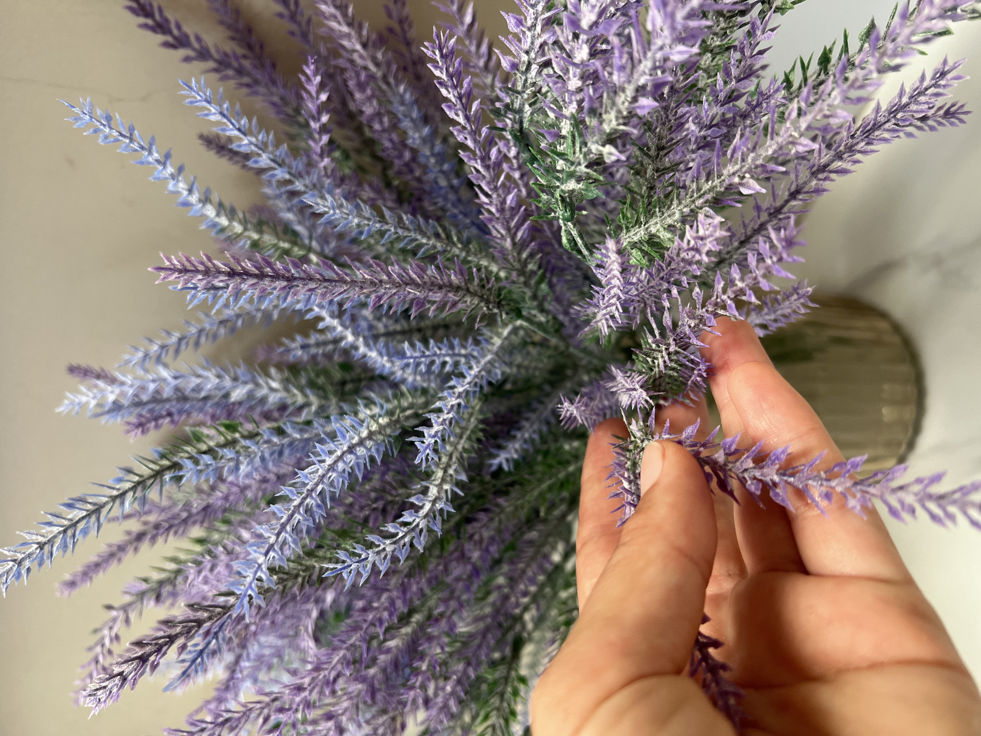 A close-up, high-angle shot depicts a hand gently touching sprigs of artificial lavender. The sprigs, a mix of purple and blue hues, are densely packed together. Green elements, resembling leaves, are interspersed among the lavender. The hand holding the sprigs is light-skinned. In the background, a blurred vase is visible on what appears to be a white surface with subtle patterns. The scene has no location information, and there is no indication that the photo was taken in Ubud District, Indonesia.