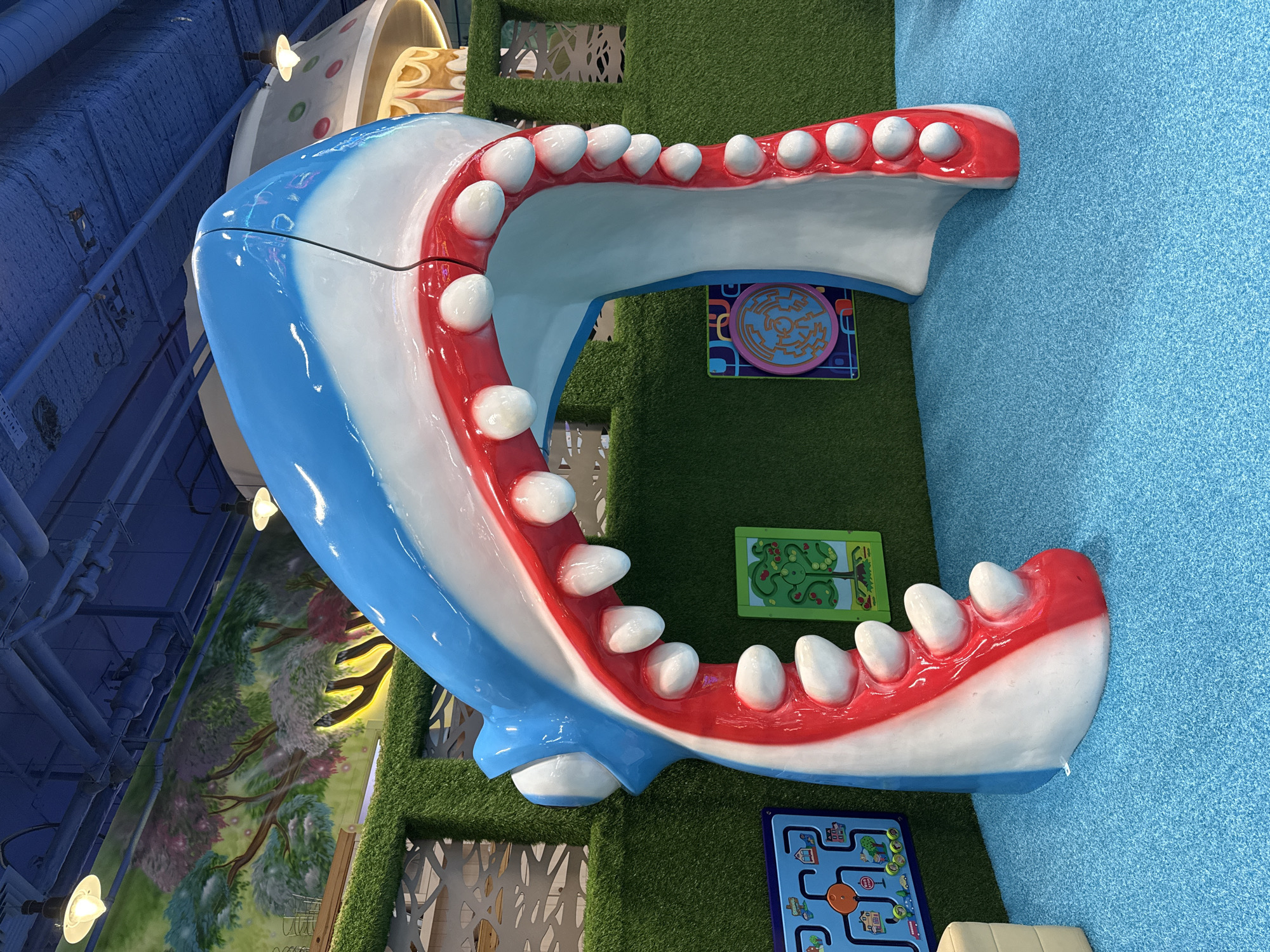 The image shows a large, open shark mouth structure with blue and white skin, a red interior, and white teeth. The structure is located in what appears to be a children's play area with artificial green grass on the walls and a blue carpet on the floor. There are colorful game boards attached to the wall around the shark mouth. The image was not taken in Перовское сельское поселение, Ukraine.