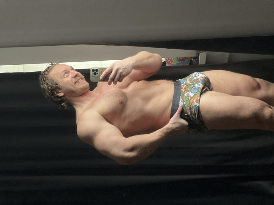 A male subject with a muscular build, curly light brown hair, and a light mustache is depicted indoors, taking a mirror selfie in Saint Petersburg, Russia. He is shirtless, displaying a well-defined torso, and wears colorful boxer briefs featuring a tropical or floral pattern with a black waistband. The subject holds a silver smartphone, identifiable by its triple-lens camera array, in his right hand to capture the reflection. His left hand is hooked into the waistband of his underwear on his left hip. He is smiling widely, with his body slightly turned and his head tilted, looking towards the camera's reflection. The reflected background reveals a dark-colored curtain on the left side of the frame, behind the subject's left shoulder, and a lighter-colored wall or surface on the right. A window sill or shelf with several small, indistinct items, including one colorful upright object, is visible in the upper right portion of the background reflection.