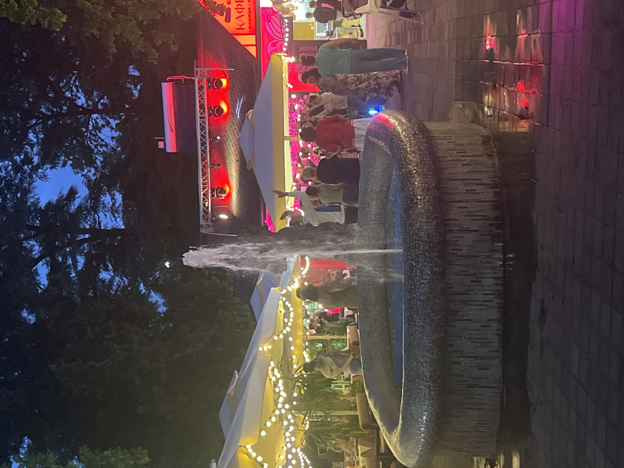 An outdoor fountain with a mosaic base is in the foreground, with water spraying upwards. In the background, a group of people are gathered in front of a stage with red lighting. Umbrellas with string lights are set up over tables, suggesting an outdoor dining or event space. The scene appears to be a lively evening gathering, possibly a cafe or restaurant in Krasnodar, Russia.