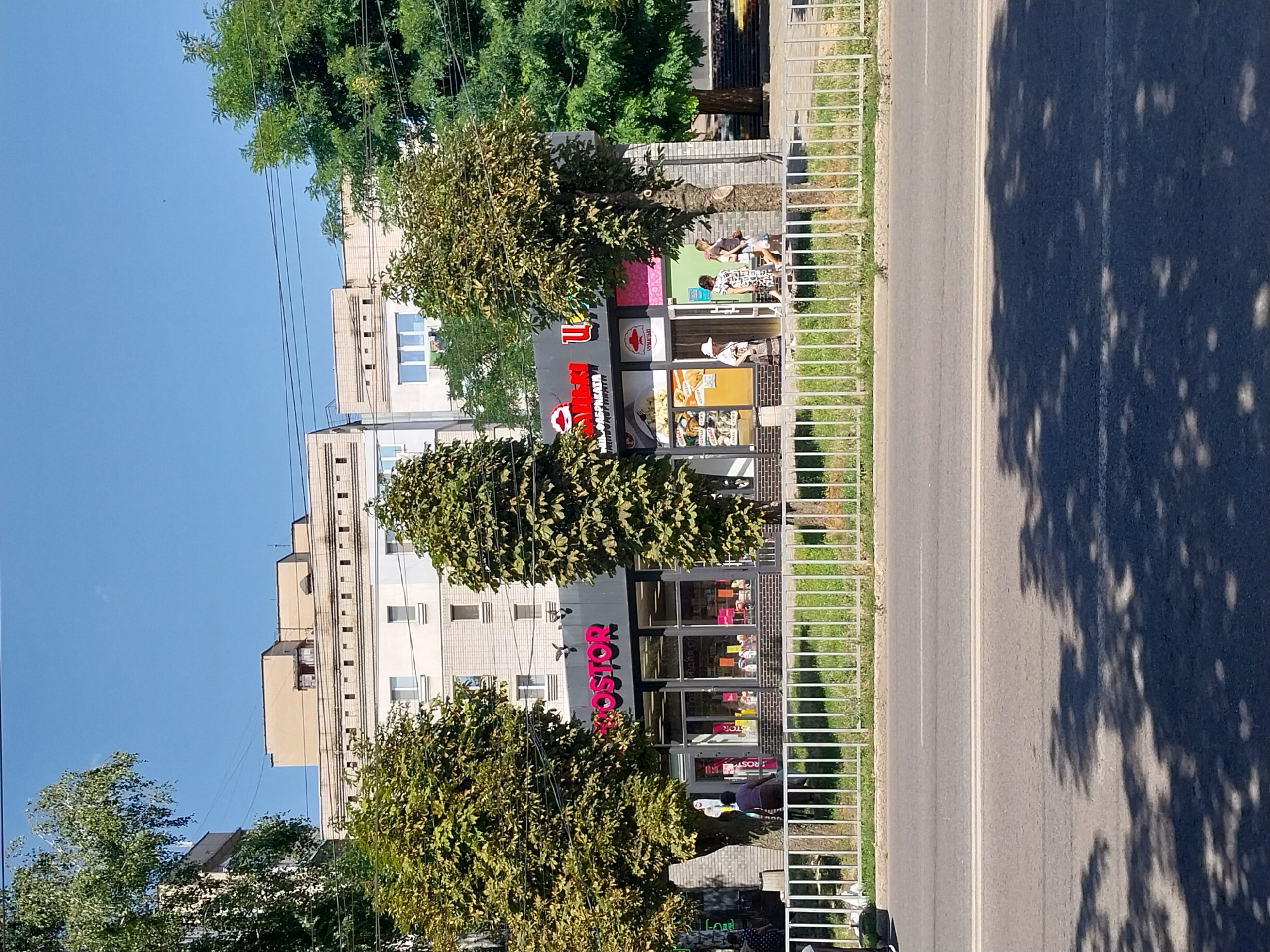 The image shows a commercial building with a bright pink sign that reads "ROSTOR". The building appears to be a store or a supermarket, with large glass windows displaying merchandise. Several trees are planted in front of the building, and a white metal fence separates the sidewalk from a street. In the background, there are apartment buildings. The scene is illuminated by bright sunlight, suggesting it is daytime. A few people are visible walking along the sidewalk. The image is taken from across the street, looking towards the commercial building. The general context suggests a typical urban street scene in Dnipro, Ukraine.