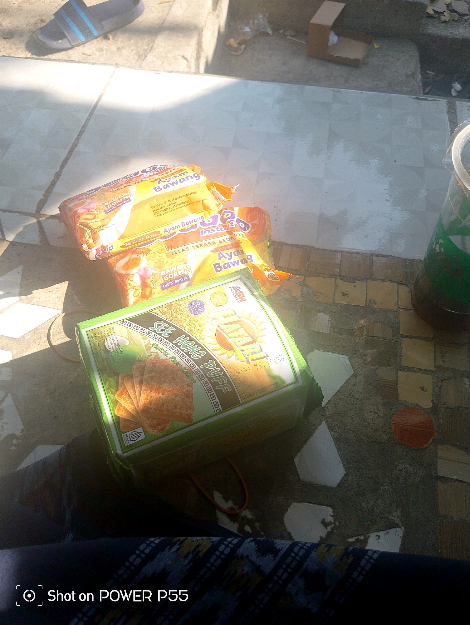In Lebaksiu, Indonesia, this image captures a collection of packaged snacks laid out on a checkered tile surface. The scene appears to be outdoors, with natural light casting shadows. Dominating the foreground is a green package of "SEE HONG PUFF" snacks, displaying an illustration of several stacked crackers. To the left of the "SEE HONG PUFF" package are two stacked, orange-and-yellow packaged instant noodle brands, "Ayam Bawang" and "Bawang Goreng." The packaging on the noodles indicates they are "Instan" and the flavor is "Ayam Bawang," which translates to "Chicken Onion." In the background, a dark liquid is contained within a clear plastic cup, and a brown cardboard box sits on a stone step. A pair of dark blue sandals with light blue straps is visible in the upper left corner, suggesting a casual, domestic setting. The overall impression is one of everyday consumables, possibly awaiting consumption or being displayed in an informal outdoor area.