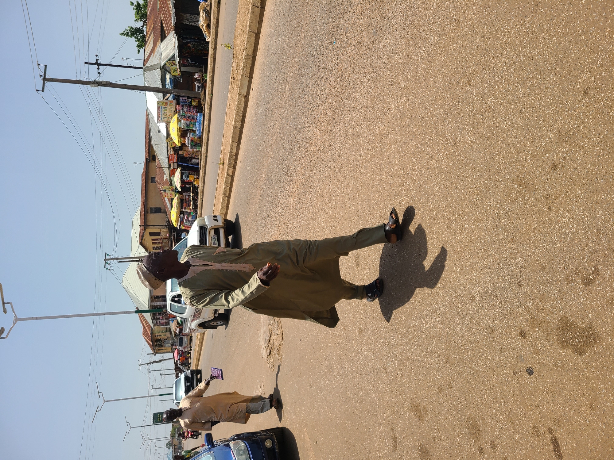 The image depicts a street scene in Bukuru, Nigeria. Two men are walking on a paved road. The man in the foreground is wearing a light green robe and pants with sandals. He is walking towards the right of the frame. The man behind him, to the left, is wearing a light brown robe and pants. He is holding a small rectangular object in his right hand and is also walking towards the right. Several vehicles are visible on the road, including a white pickup truck and a blue car. In the background, there are buildings, stalls with umbrellas, and utility poles with overhead wires. The sky is clear and the scene appears to be well-lit by sunlight, casting shadows on the road.