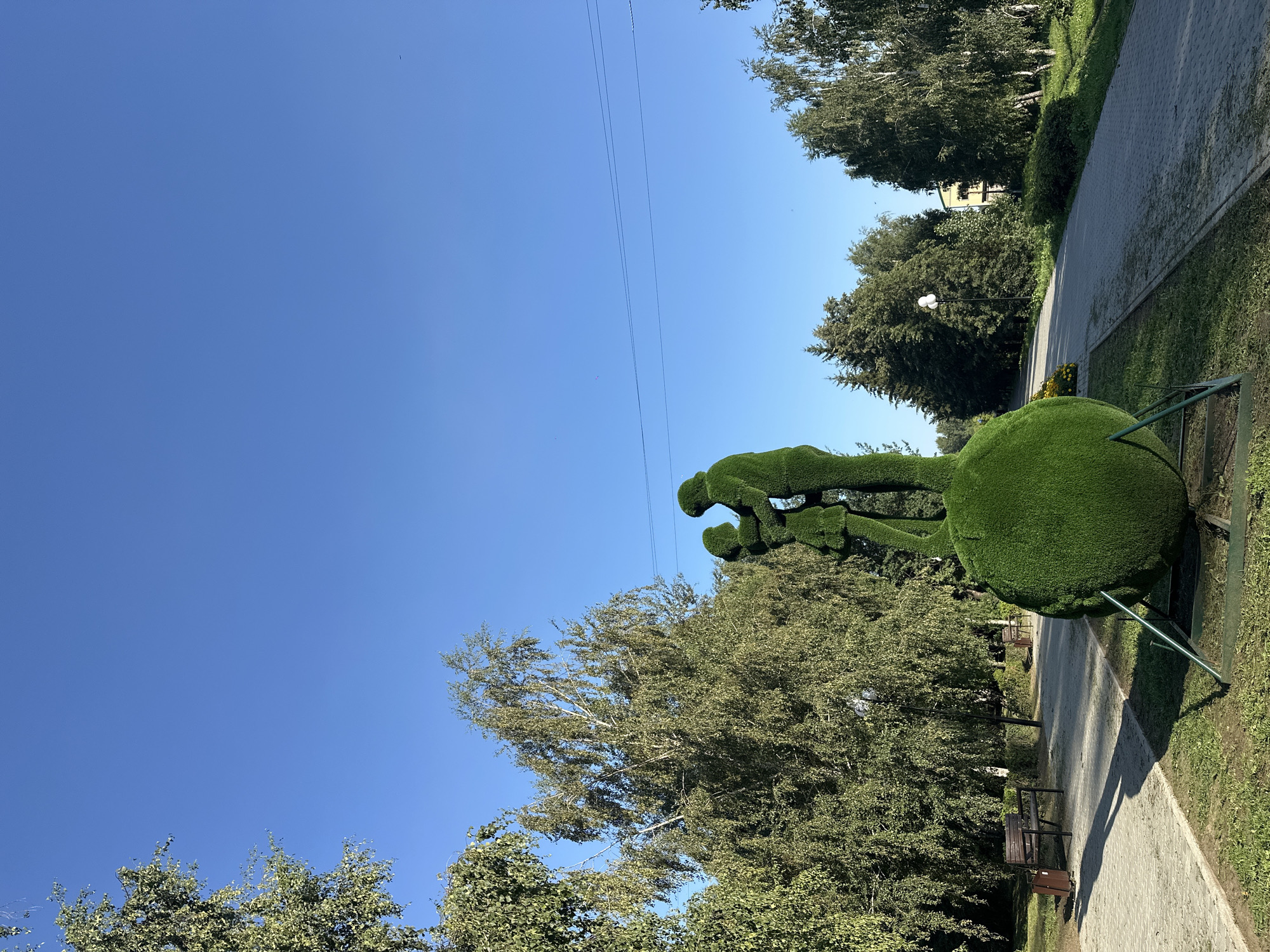 An outdoor, low-angle, full shot captures a topiary sculpture of two figures embracing, situated in the city of Nogornyi, Russia. The sculpture, made of green artificial grass, depicts a man and a woman, with the woman being smaller than the man. They stand on a large, round, green base, supported by a metal frame. The figures are silhouetted against a clear, bright blue sky, with the sun casting long shadows to the left.
The background consists of a paved walkway curving to the right, lined with trees and benches. The trees are predominantly deciduous with green foliage, with some evergreen trees visible further back. A lamppost stands in the midground to the left of the walkway. In the distance, a yellow building with a grey roof can be seen through the trees. The environment appears to be a park or public garden. The overall lighting suggests it is daytime, with the sun positioned high in the sky.
