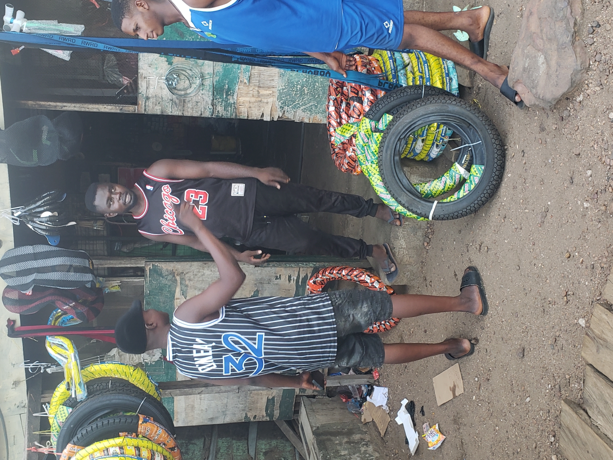 The image depicts an outdoor market scene in Lokoja, Nigeria, during daylight hours. Three young men are gathered around a tire stall. The ground is unpaved, a mix of dirt and gravel, with a large rock visible on the right.
One man, wearing a blue tank top with "RIGORE" visible, stands on the right side of the frame, holding a blue strap printed repeatedly with "RWRD" text. Beside him are numerous motorcycle or bicycle tires, stacked and wrapped in colorful plastic packaging. Some wrappings are yellow and green, featuring text like "TUDOS" and "TUDOS TYRE", while others are orange with "TENTOR". A larger, dark tire with a visible tread pattern is also prominently displayed.
Inside a rustic, wooden stall or shop area, another man is seen wearing a black Chicago Bulls jersey with the number "23". He looks towards the viewer with a neutral expression. The third man, donning a black and white striped jersey with "RNEW" and the number "32" on it, and a black cap, stands facing away from the camera, interacting with something at his side.
The setting suggests a casual, bustling commercial environment typical of a street market, with various miscellaneous items like hangers, discarded cardboard, and snack wrappers scattered around. The bright, even lighting indicates it is daytime. The men are dressed in casual jerseys and sandals, suitable for a warm climate.