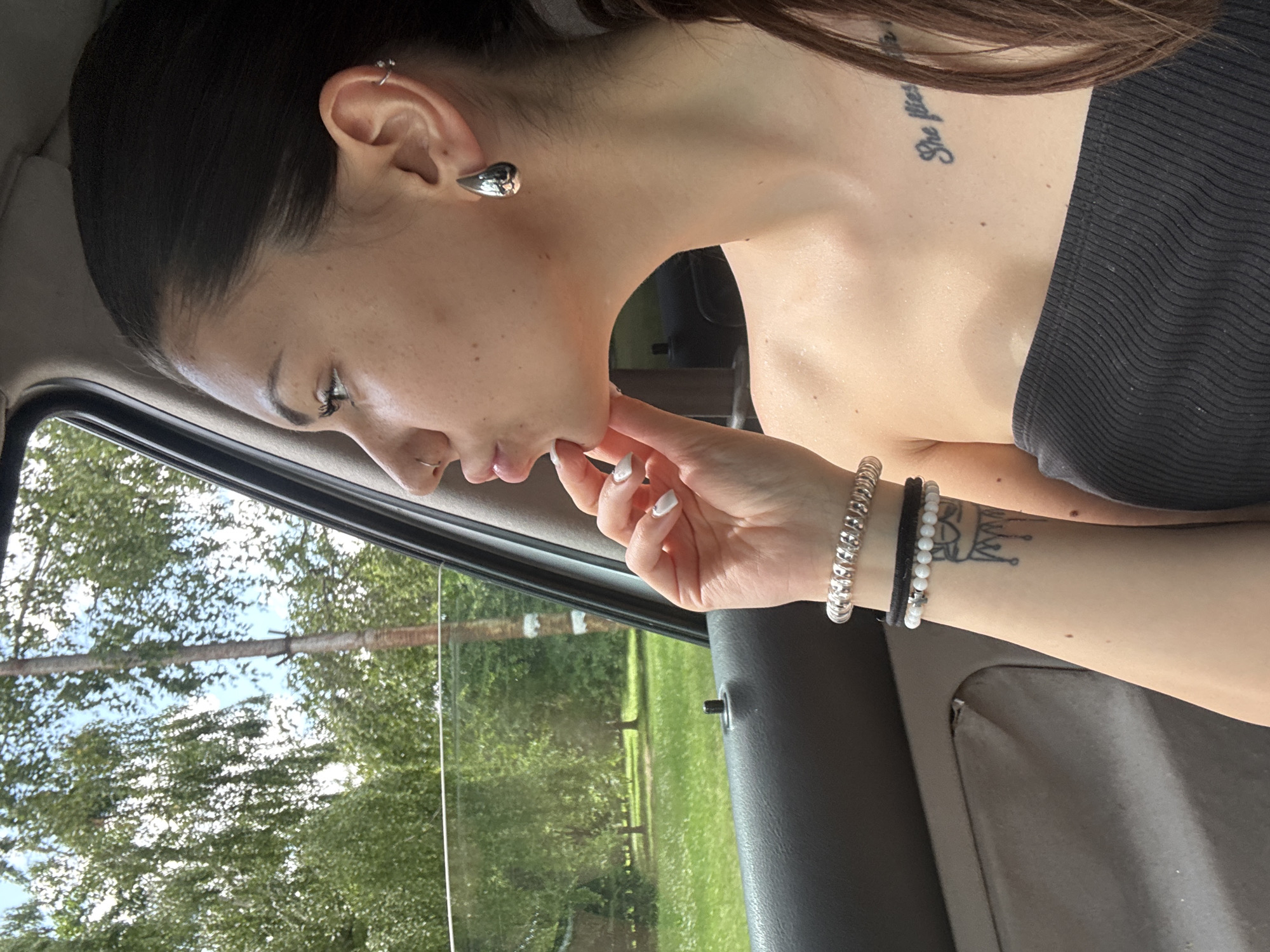 The image features a young female observed in profile within a vehicle, looking towards the right out of a partially open window. Her dark hair is pulled back, with some strands visible cascading over her shoulder. Her skin tone is fair, displaying minor freckles across her nose and cheek area. She has a small silver hoop nose ring in her left nostril. Her visible left ear is adorned with a large, polished silver, teardrop-shaped earring in the lobe, a small silver hoop piercing in the upper cartilage (helix), and another small silver ring piercing likely in the conch or flat area.
Her left hand is raised, with fingers lightly touching her chin or jawline; her fingernails are painted white. On her left wrist, she wears a spiraled silver-toned metal bracelet alongside a stack of smaller bracelets, including a strand of white beads and a black elastic band with a small silver cross charm. A tattoo resembling a crown with dripping elements is visible on her inner left forearm. On her upper left chest, a text tattoo begins with the words "She flies...". She is wearing a black, ribbed, strapless top.
The car's interior is visible as a dark gray door panel, featuring a visible door lock knob. Outside the vehicle, a bright, sunny daytime scene unfolds with a lush green environment. Numerous deciduous trees with full foliage dominate the background, suggesting summer. Two distinct tree trunks are visible, one slender and light-colored with horizontal markings, and another thicker, darker trunk. The ground is covered in green grass. Patches of blue sky with some white clouds are discernible through the tree canopy. A faint, dark, indistinct shape is visible in the distant background, possibly a structure or another object. The location is in the city of Sofia, Bulgaria.
