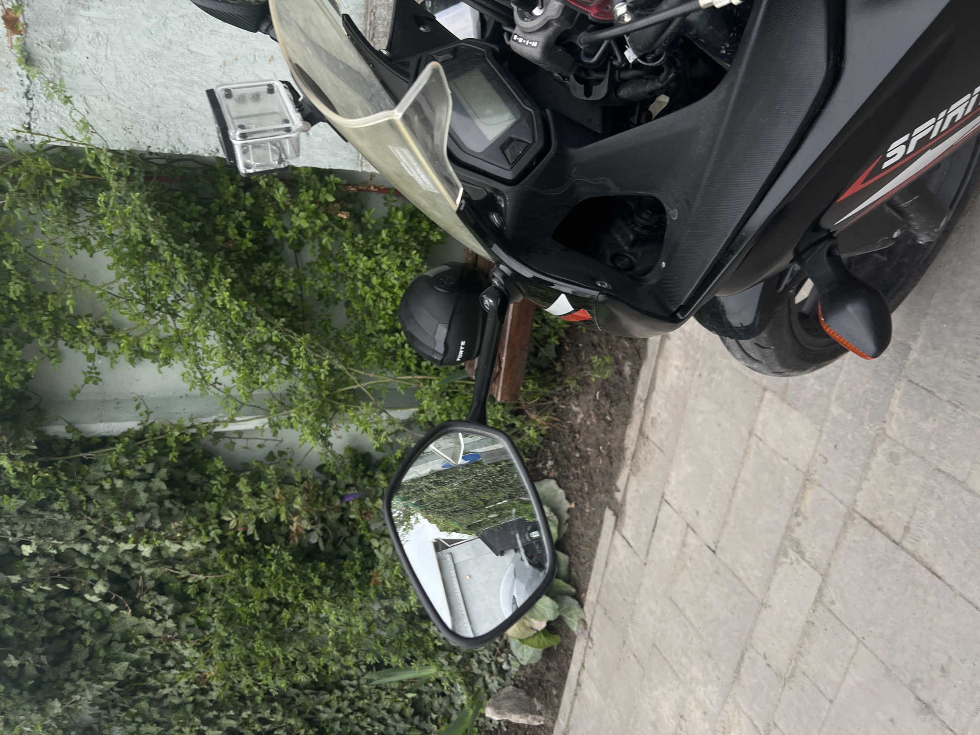 The image shows the front section of a black motorcycle parked on a paved surface, possibly a sidewalk or driveway, next to a wall covered in dense green foliage. The motorcycle's side mirror reflects a building and vegetation, providing a sense of the surroundings. A black motorcycle helmet is positioned near the handlebars. Based on the request to include the location in the city of Horodok, Ukraine, the motorcycle is located there. The image captures a static scene of the motorcycle in its environment. No people or actions are visible in the frame.