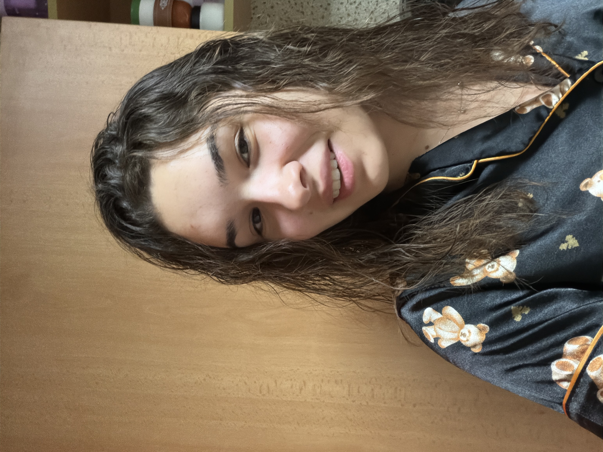 A person with wet, dark, wavy hair is smiling and looking at the camera. They are wearing a dark-colored satin pajama top with a yellow trim and a repeating pattern of teddy bears. The background is a light brown, possibly a wooden surface. There is no indication of location or city. The scene appears to be indoors.