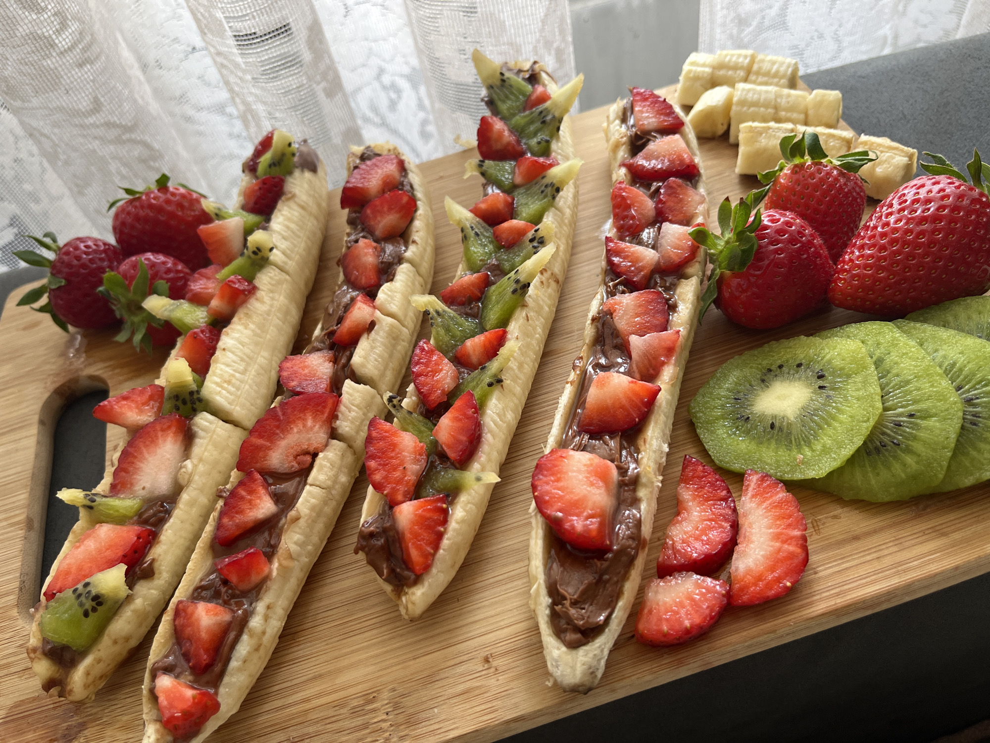 An overhead shot showcases a vibrant fruit and chocolate dessert arrangement laid out on a light-colored wooden cutting board, likely indoors in Sofia, Bulgaria. The main subjects are multiple "banana boats," created by slicing bananas lengthwise and filling them with a rich chocolate spread, chopped red strawberries, and sliced green kiwi pieces. Some of these banana boats also feature smaller banana chunks.
To the left of the board, a cluster of whole, ripe red strawberries rests, while the bottom right features neatly fanned slices of kiwi. Further up on the right side, more whole strawberries and additional small, light-colored fruit pieces (possibly more banana) are visible. The arrangement suggests a freshly prepared snack or dessert, emphasizing natural sweetness and color.
The background reveals a sheer white curtain with a subtle pattern, suggesting a window and natural light illuminating the scene, likely during daytime. A dark, possibly grey or black, surface serves as the tabletop underneath the cutting board. The lighting is soft and even, highlighting the textures and fresh appearance of the fruit and chocolate. No text is visible in the image. The overall impression is one of a healthy yet indulgent treat, artfully presented.