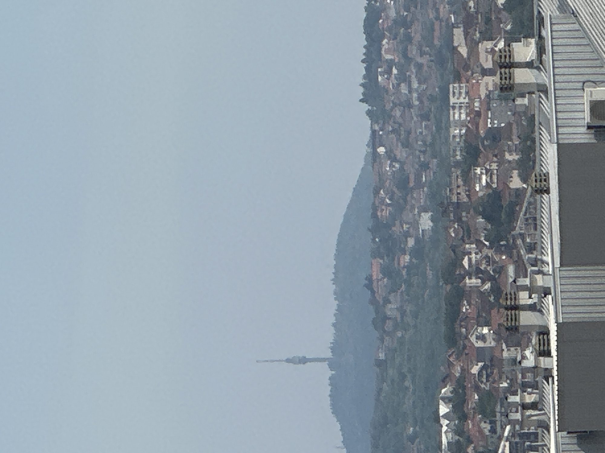 The image captures a distant view of Belgrade, Serbia, focusing on a sprawling residential area nestled at the foot of a hill. Dominating the skyline on the hill's crest is the Avala Tower, a prominent telecommunications tower. The foreground shows the rooftops of several modern buildings, characterized by grey, corrugated metal surfaces and numerous chimneys. Beyond these buildings, the mid-ground reveals a dense collection of houses with terracotta roofs, characteristic of residential neighborhoods. The background features the undulating terrain of the hill, heavily wooded, with the Avala Tower standing tall and slender against a hazy, pale blue sky. The overall impression is of a hazy, daytime cityscape with a mix of urban development and natural landscape.