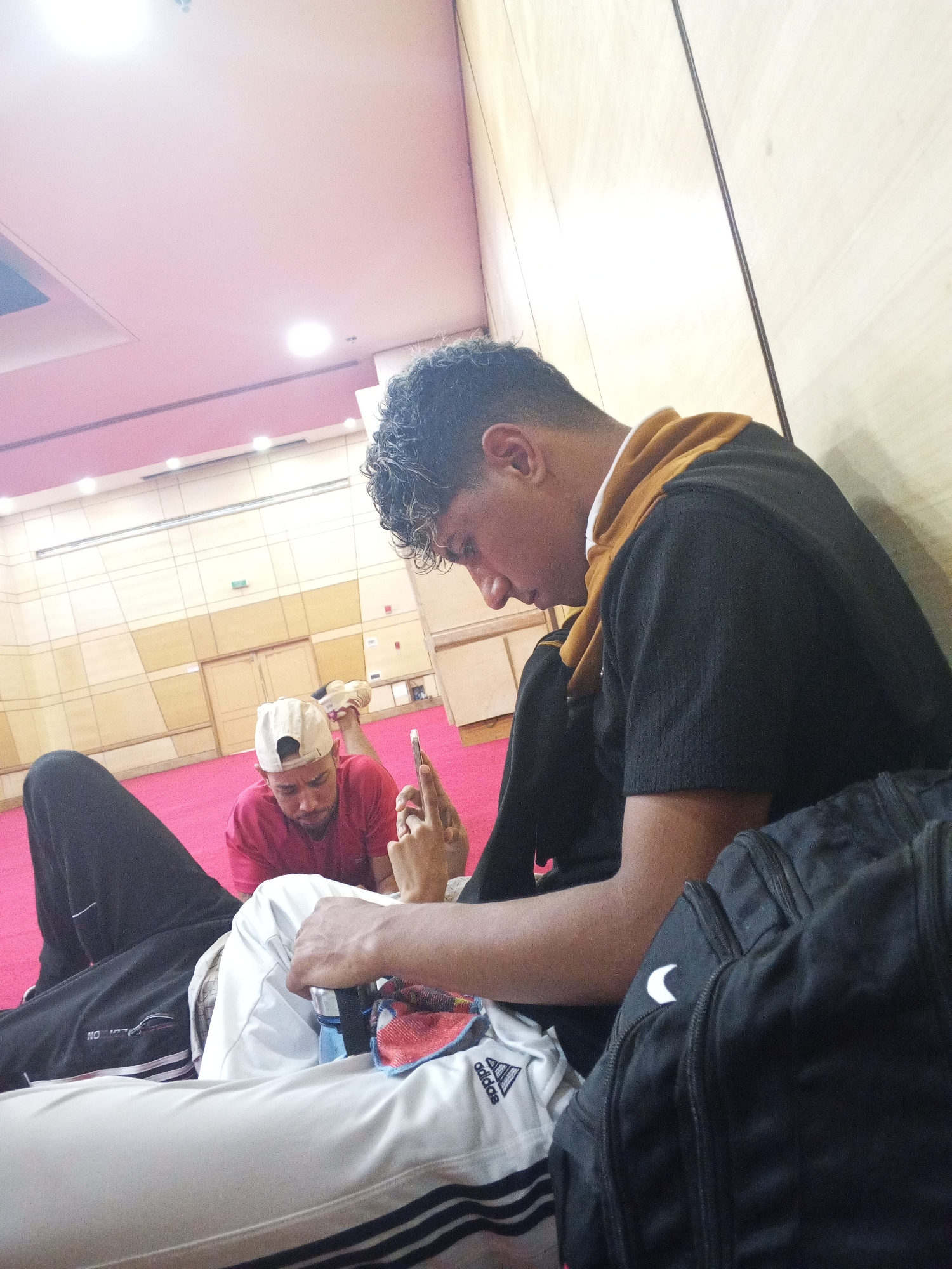 Three young men are sitting on the floor indoors. The building appears to be an institutional hall or conference center, with tiled walls and a pink ceiling. The floor is carpeted in red.
The man in the foreground is sitting with his back against the wall, wearing black athletic pants, a white Adidas track jacket, a black sweatshirt and a mustard-colored undershirt. Next to him is a black Nike backpack. A second man, wearing a red shirt and white baseball cap, is lying on the floor behind him, holding a cell phone up in his right hand.
It is impossible to determine the location in the city of Porlamar, Venezuela from this image.