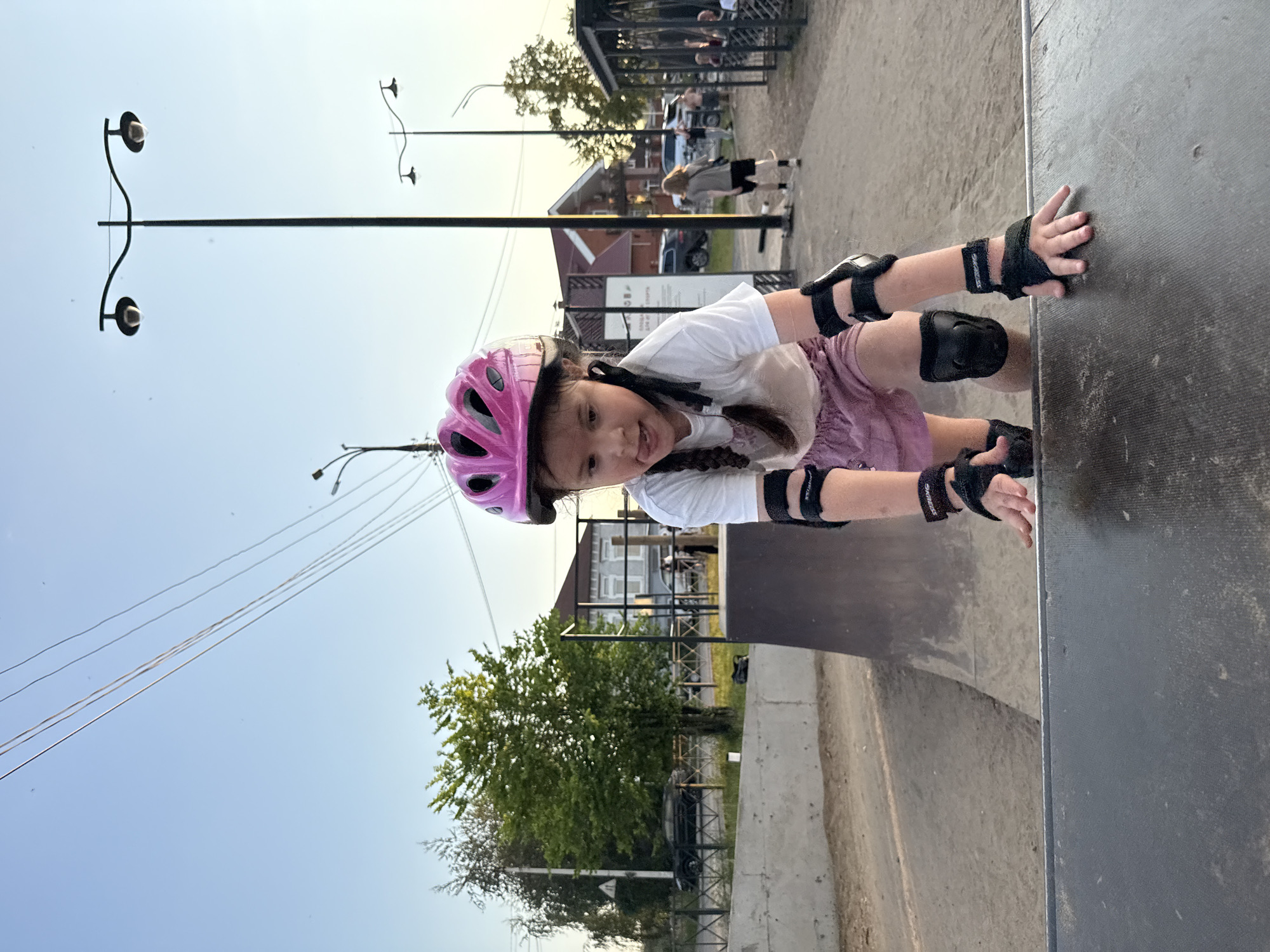 A young girl wearing a pink helmet and protective gear is on a skateboard ramp. She is wearing a white t-shirt, a pink skirt, elbow pads, and knee pads. Her hands are on the ramp, and she is looking at the camera with a playful expression, sticking her tongue out slightly. In the background, there is a skate park with other people, trees, and buildings. The scene appears to be set in a park or outdoor recreational area. The lighting suggests it is daytime, likely late afternoon or early evening, given the soft light. The sky is clear and blue. There are streetlights visible in the upper left corner of the image. Visible text includes brand names on the protective gear. The overall mood is one of fun and activity.