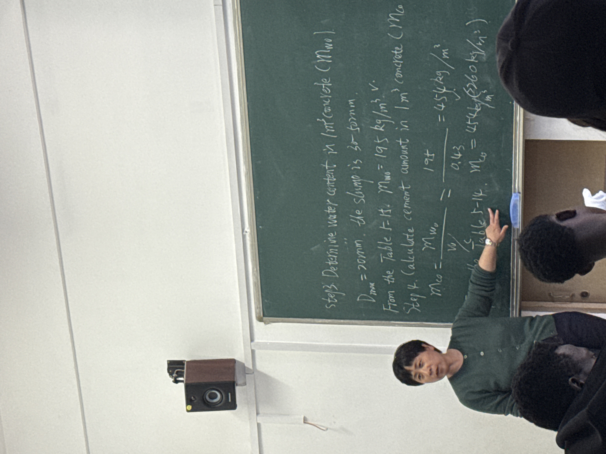 The image captures an indoor classroom scene featuring a woman, likely a teacher, instructing students in front of a green chalkboard. The teacher, dressed in a green long-sleeved top, is actively pointing at the board with her right hand and appears to be speaking. Her dark hair is cut short. Several students, with dark, curly hair, are partially visible in the foreground and to the right, mostly seen from behind, suggesting they are seated and facing the chalkboard.
On the left side of the white wall, a dark speaker with a visible yellow light is mounted. The classroom has white walls and a white ceiling.
The chalkboard is filled with handwritten text and calculations related to concrete. The visible text outlines steps for determining concrete mix components:
"step3. Determine water content in 1m² concrete (Mwo).
Dmax = 20mm, the slump is 30-50mm.
From the Table 5-15. Mwo = 195 kg/m³. v.
Step 4. Calculate cement amount in 1m³ concrete (Mco)
Mco = Mwo / (W/C)
= 195 / 0.43
= 454 kg/m²
(fbe5-14. Mco = 454 kg/m² or 560 kg/m²)"
The scene depicts an educational activity, likely a lecture or problem-solving session in a technical or engineering subject.