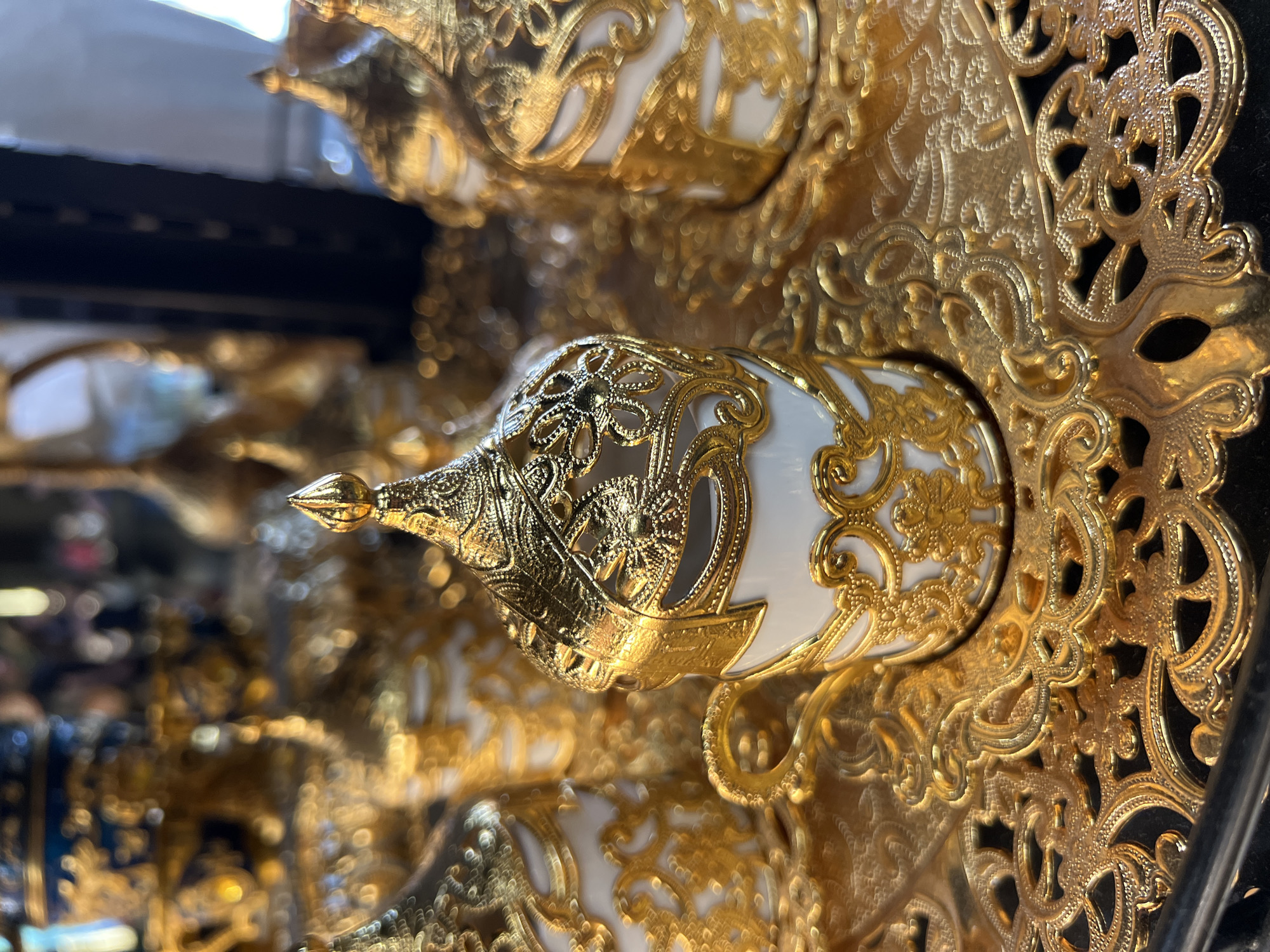 The image presents a close-up of an ornate, gold-colored cup, likely a demitasse, set against a blurred background of similar items. The cup features a white porcelain or ceramic interior adorned with gold patterns, encased by intricate gold filigree work that forms detailed floral and scroll motifs. A prominent, pointed finial or lid top, also in gold, extends from one end of the cup. The main subject rests on, or is adjacent to, a base or saucer equally embellished with elaborate, cut-out gold patterns.
The scene appears to be an indoor display, possibly within a shop, market, or exhibition in Krasnodar, Russia, given the multitude of similar, highly decorative items visible out of focus in the background. The lighting emphasizes the reflective qualities of the gold, highlighting the meticulous craftsmanship and luxurious design of the artifacts. No human activity or individuals are depicted; the focus is solely on the static, artistic objects.
The style of the intricate gold filigree and the overall opulent aesthetic suggest a traditional design influence, potentially from Ottoman, Arabian, or Eastern European cultures, fitting the geographical and historical context of Krasnodar. The objects evoke a sense of grandeur, historical richness, and fine artistry. There is no visible text in the image.