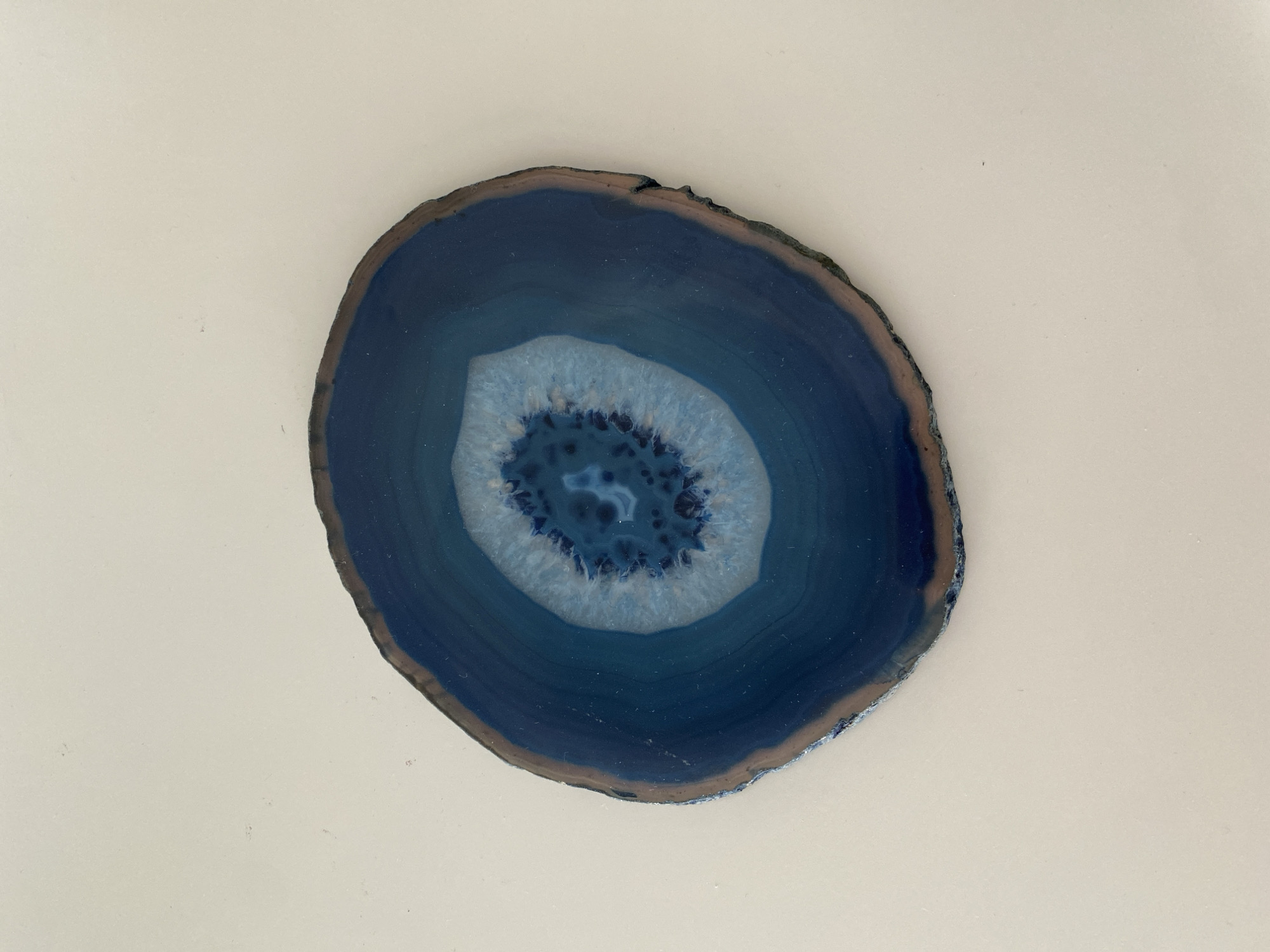 The image features a polished slice of blue agate, centered on a plain, off-white surface. The agate exhibits concentric bands of varying shades of blue, with a lighter, nearly white central core that has a crystalline structure. The outer edge of the agate has a rough, unpolished rim. The subject is well-lit and in focus, suggesting a static setting. The image offers no information about the city of Votkinsk, Russia. The surface the agate is resting on appears smooth and featureless.