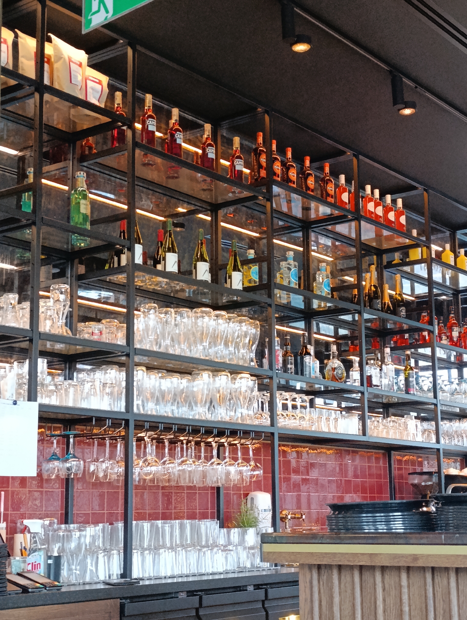 The image depicts a well-stocked bar shelf in a commercial establishment, likely a restaurant or pub, located in Mysiadło, Poland. The shelves are made of black metal and are filled with various bottles of liquor, wine, and other beverages. Several rows of glassware, including wine glasses and tumblers, are neatly stacked on lower shelves. The background features a red tiled wall. The lighting in the establishment appears to be ambient, with some spotlights casting a warm glow. The overall impression is one of a clean and organized bar area, ready for service. No people are visible in the image.
