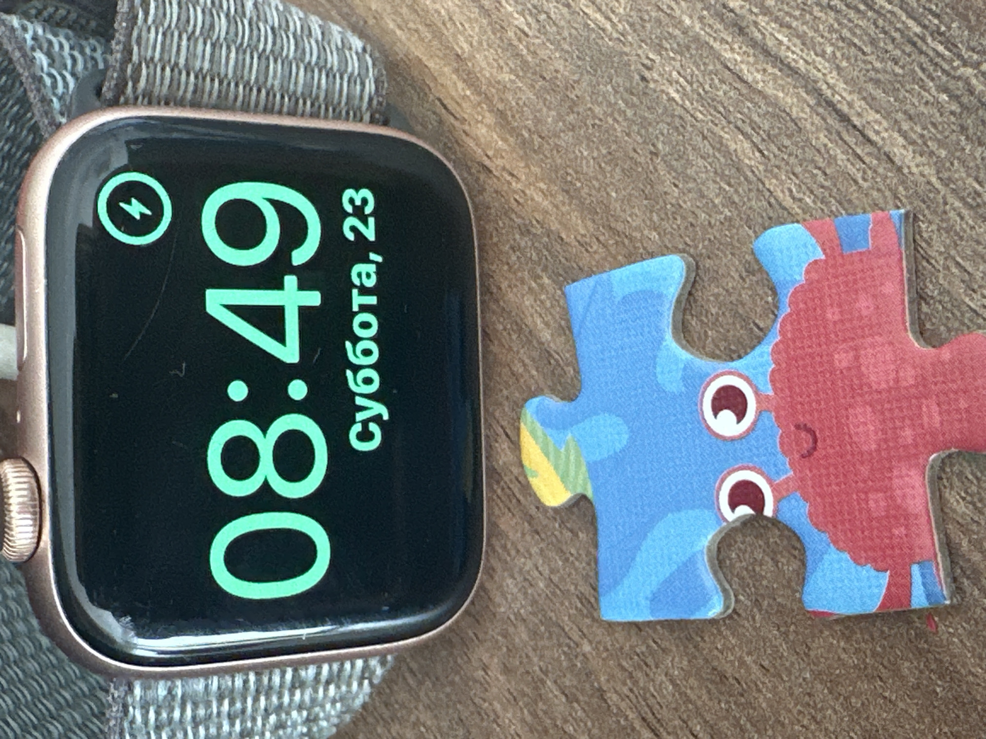 A close-up shot on a wooden surface features an Apple Watch and a jigsaw puzzle piece. The Apple Watch displays the time as 08:49, with a charging symbol visible in the top left corner. The date displayed is "суббота, 23." To the right of the watch, there is a single jigsaw puzzle piece depicting cartoonish sea creatures, with a blue and red color scheme. No people are visible in the image. The setting is undefined but does not appear to be in Fethiye, Turkey.