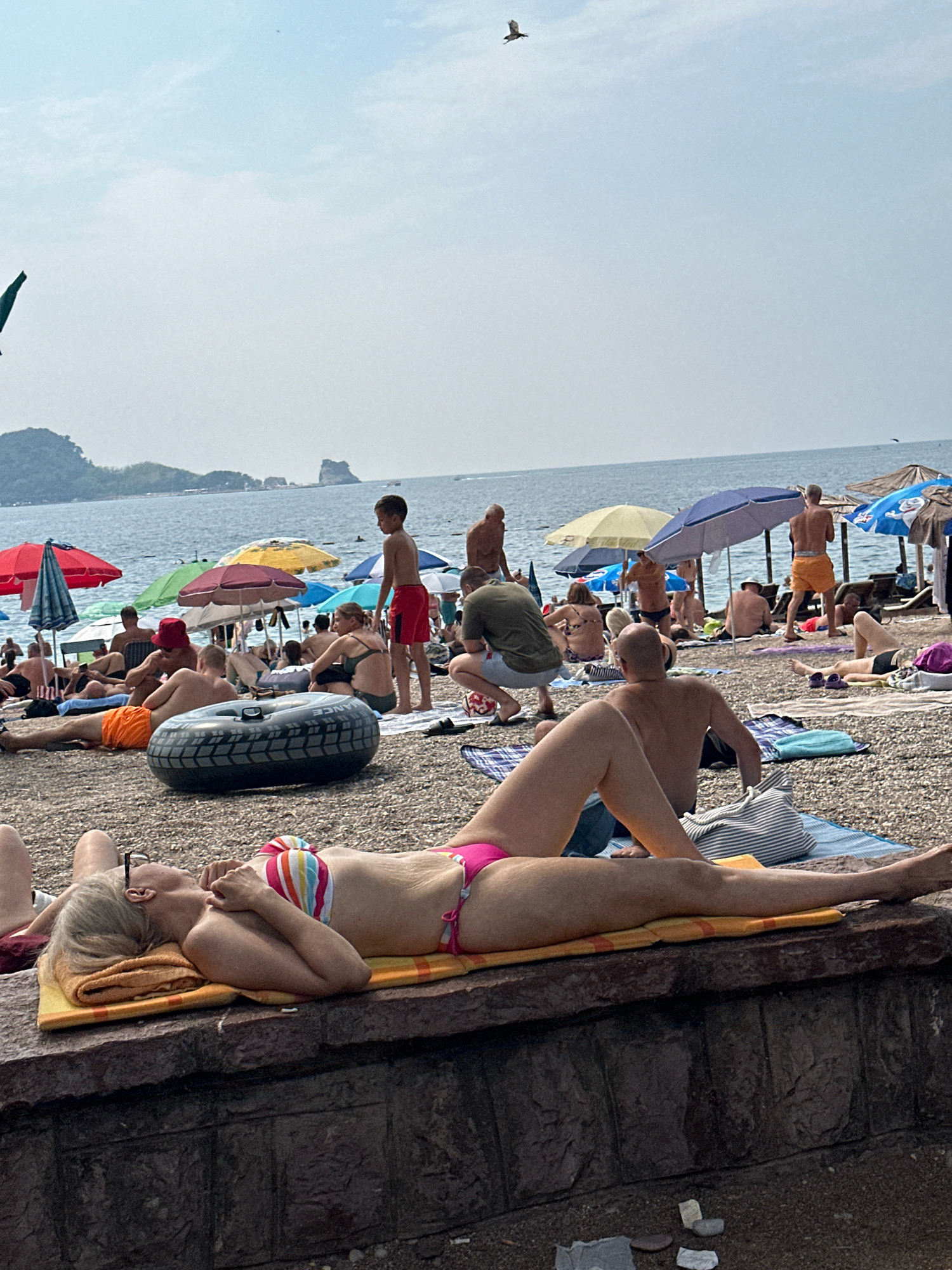 The image shows a crowded beach scene on a sunny day, likely in Budva, Montenegro, given the characteristic rocky coastline and mountainous backdrop. In the foreground, a woman with blonde hair, wearing glasses and a colorful striped bikini, is sunbathing on a yellow towel laid on a stone wall. Her eyes are closed, and her hands are resting on her chest. Behind her, partially visible, are the legs of another person.
The beach itself is covered in pebbles, and numerous people are scattered across it, many sunbathing on towels or beach chairs. Beach umbrellas, in various colors including red, yellow, and blue, provide shade. In the middle ground, an inflatable black tire-shaped object is lying on the pebbles. Several individuals are visible in the background, some standing, some sitting, and others lying down. A young boy in red shorts stands near a blue umbrella. The sea stretches out in the background, with a hazy mountainous landscape on the horizon. The sky is bright and partially cloudy, with a bird soaring in the distance.