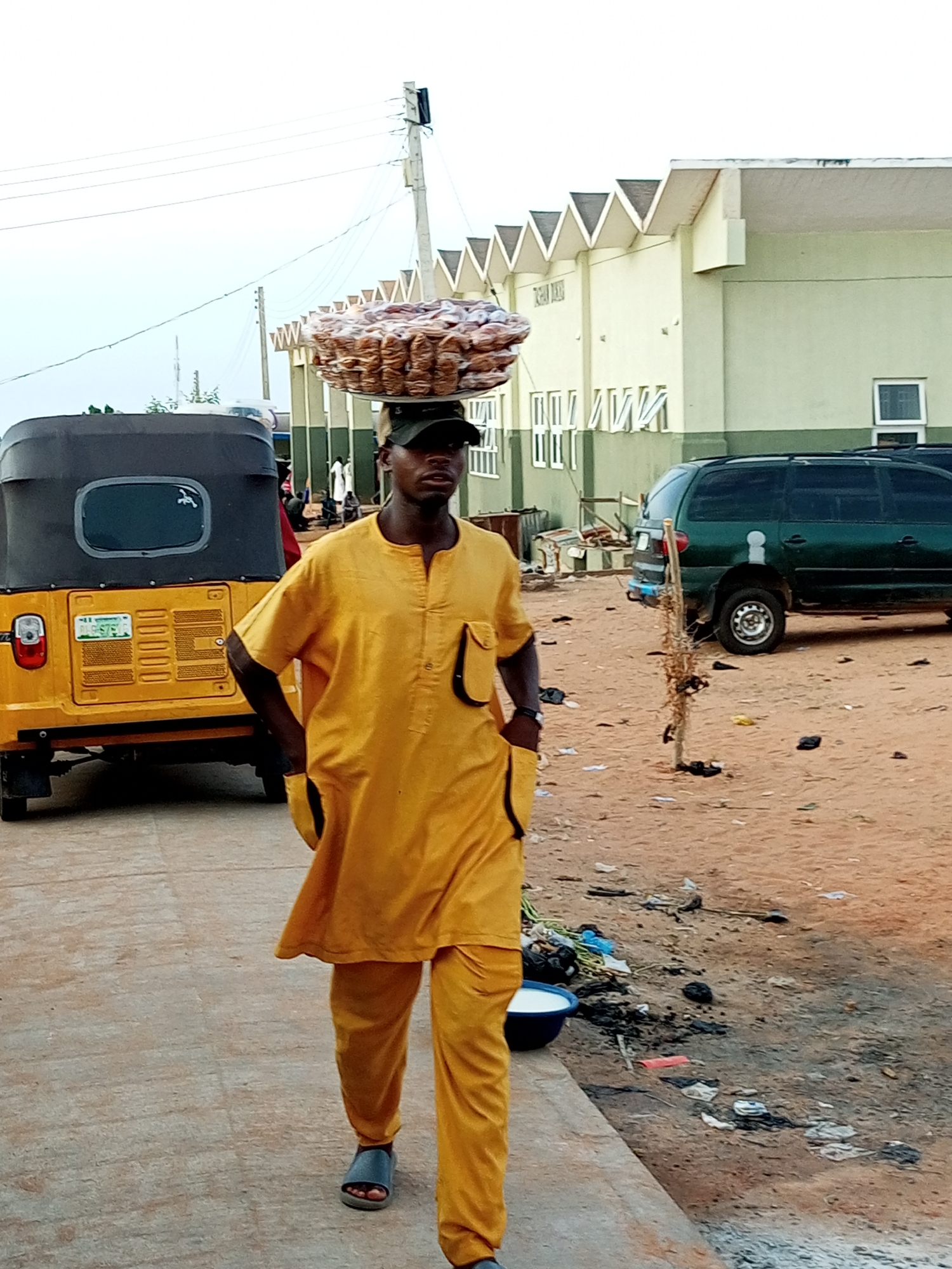 A man in a yellow traditional outfit and a black cap walks with a large circular tray of packaged goods balanced on his head. He is on a paved walkway. To his left is a yellow three-wheeled auto-rickshaw. In the background, a green SUV is parked next to a light-colored building with a distinctive zigzag roofline. The ground is sandy and littered with debris. This scene is in Gombe, Nigeria.