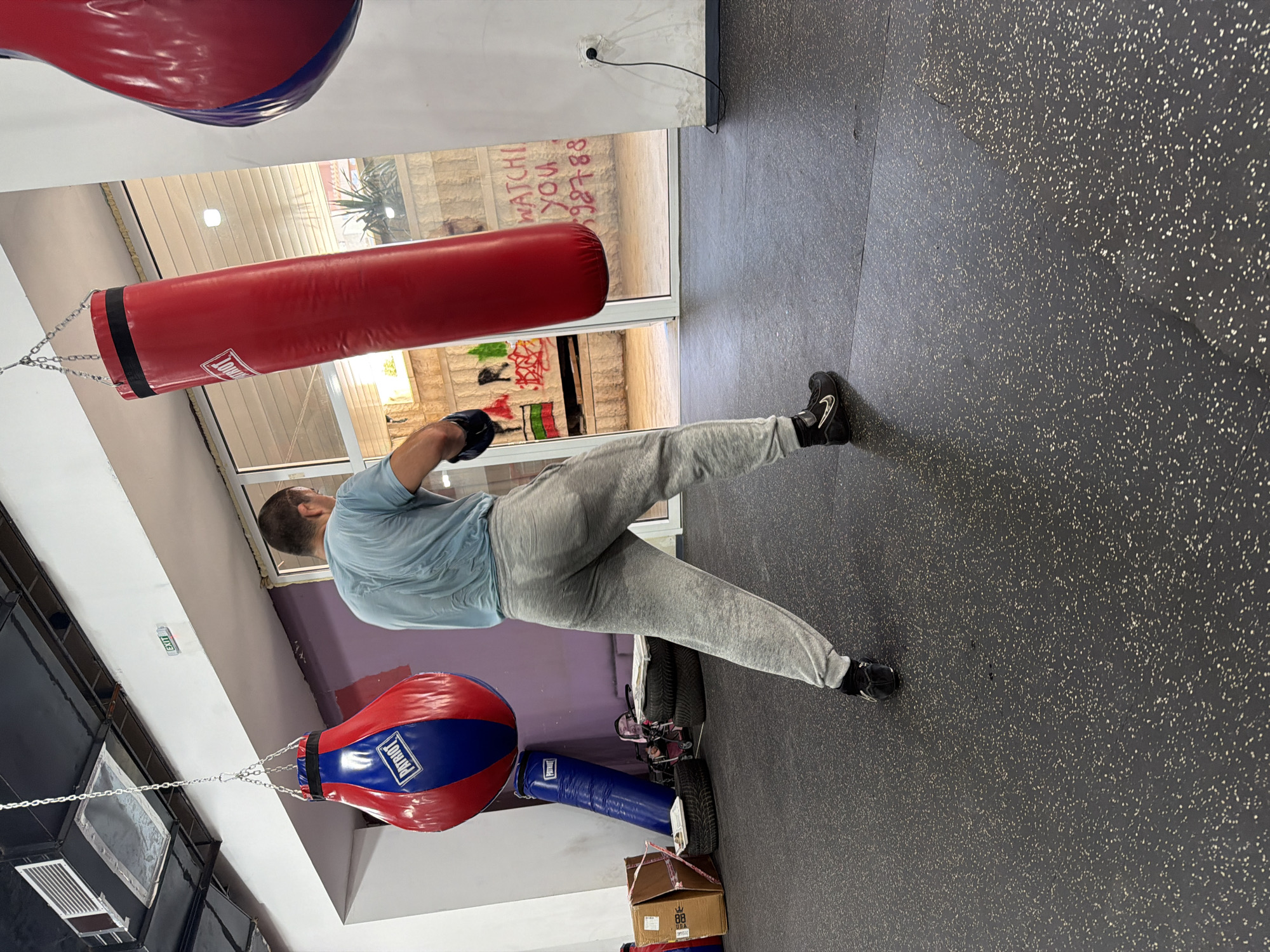 A male boxer is training in a gym in Blagoevgrad, Bulgaria. He is wearing a light blue t-shirt and grey sweatpants. He is wearing boxing gloves and appears to be shadowboxing or striking a heavy bag. The gym has several punching bags, including a large red one and a smaller red and blue one. The floor is made of dark rubber matting with white speckles. In the background, there is a window with graffiti on it, and some exercise equipment. The lighting in the gym is overhead fluorescent lighting.