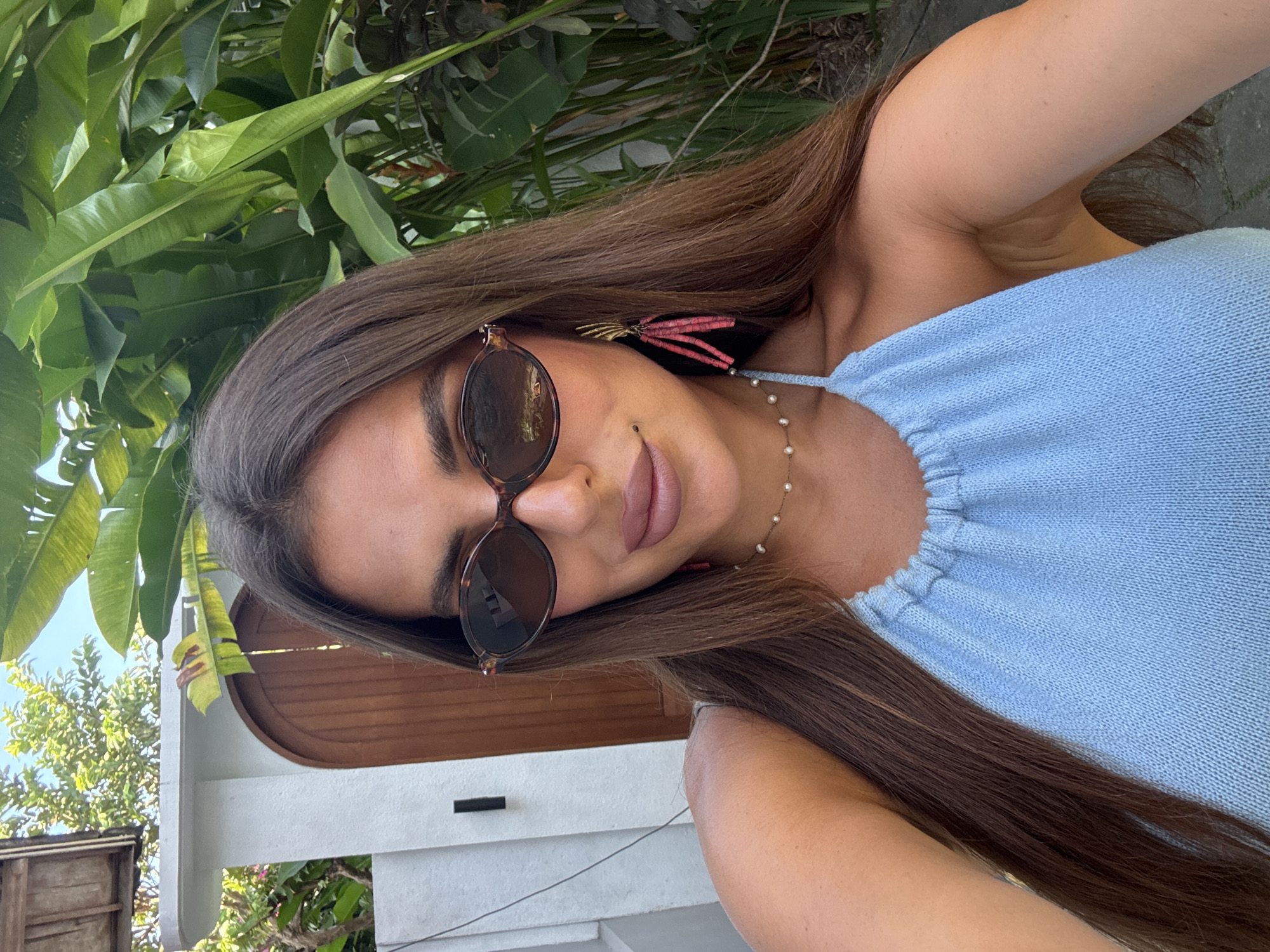 A woman is posing for a selfie in an outdoor location that appears to be in a tropical climate, likely near North Kuta, Indonesia. She has long brown hair, is wearing sunglasses, a light blue top, and a beaded necklace. Green foliage and a building with a wooden door are visible in the background.