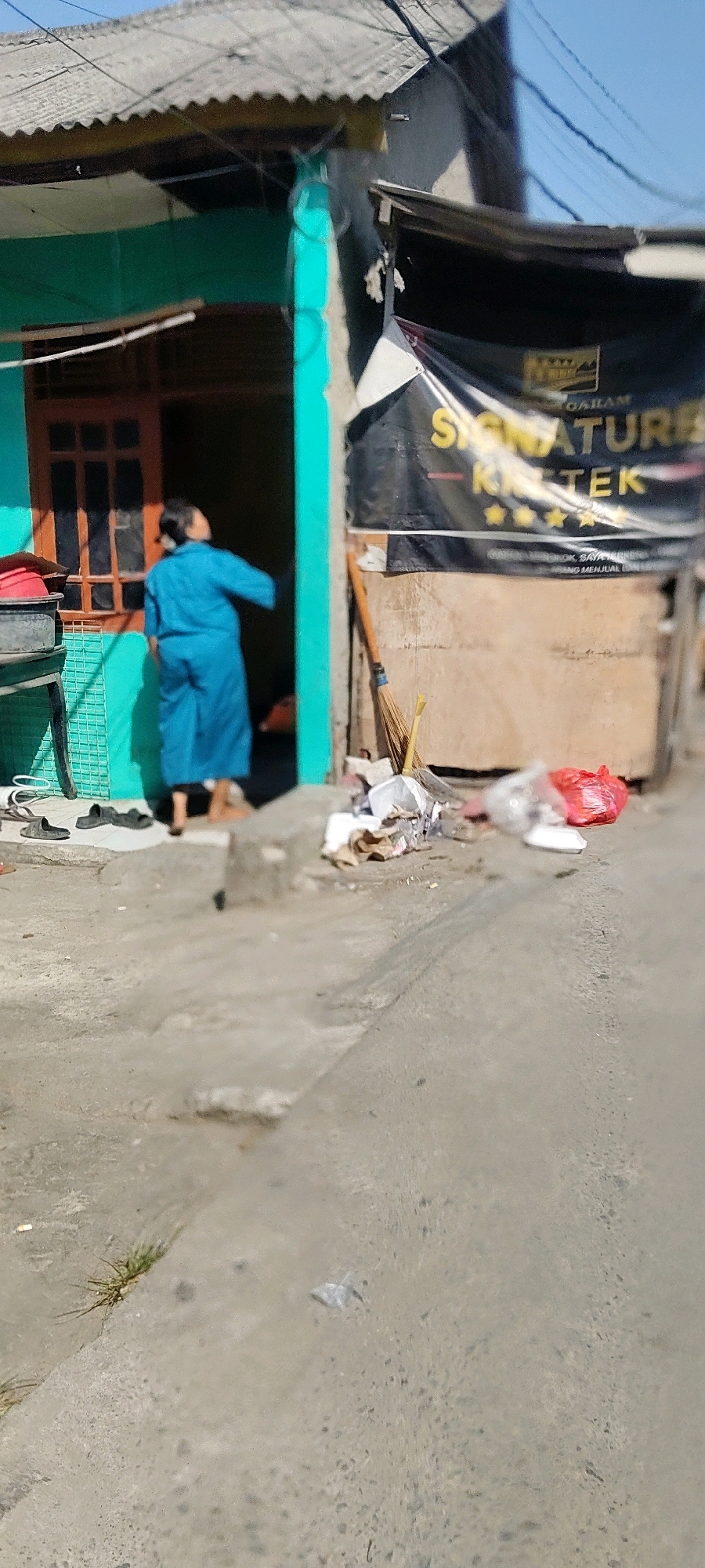 The image depicts an outdoor scene in the city of Mekarmukti, Indonesia. A person, appearing to be of adult age and wearing a blue robe or dress, is standing in the doorway of a teal-colored building with a brown-framed window. The person is facing away from the camera and has their head tilted upwards. In front of the building, to the right of the entrance, a broom is propped against a wooden structure, and there is a pile of assorted trash including plastic bags, one of which is red, and various paper and other debris. Two pairs of black sandals are placed on the concrete ground to the left of the building entrance. The building itself has a corrugated metal roof and appears to have a banner with text in yellow and black stretched across the front, partially obscuring the wooden structure. The ground in the foreground is a rough concrete surface with some patches of dry grass and small debris. The overall impression is that of a modest urban or semi-urban environment under a clear sky.