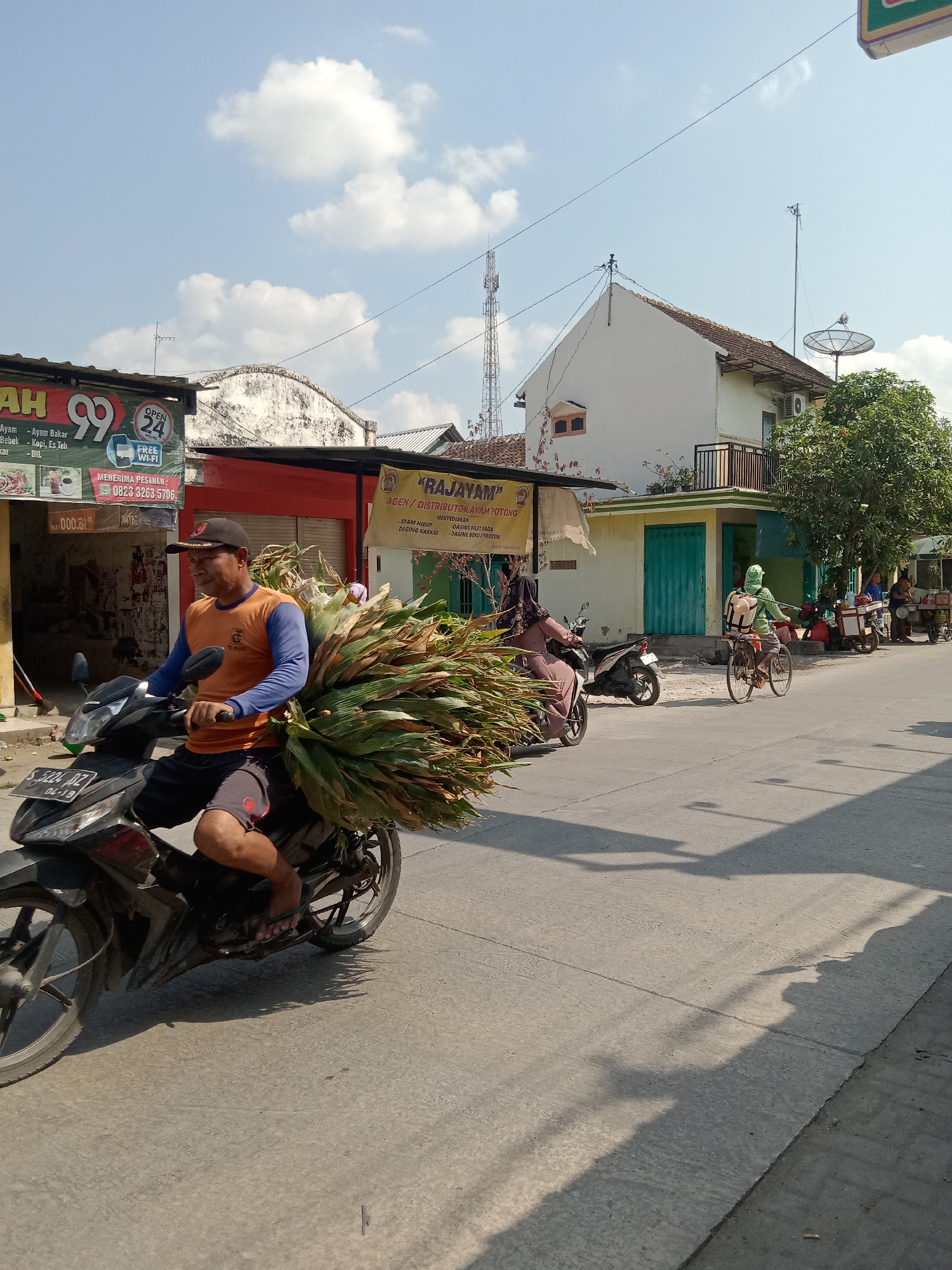 In the city of Klepek, Indonesia, on a sunny day, a man is riding a motorcycle carrying a large load of palm leaves. He is wearing an orange shirt with blue sleeves, black shorts, and a black baseball cap. The motorcycle is black, and the license plate reads "S 5224 DZ 04-19". The man is on a paved road, with buildings and shops lining the street. To the left of the motorcycle is a shop with a sign that says "99" and offers "OPEN 24 HOURS" with "FREE WI-FI". To the right of the motorcycle, a person on a bicycle wearing a green jacket and carrying a backpack is also on the road. Another person on a motorcycle is seen further down the street, as well as two individuals standing near a cart. The sky is blue with scattered white clouds. The environment suggests a commercial area with ongoing daily activities.