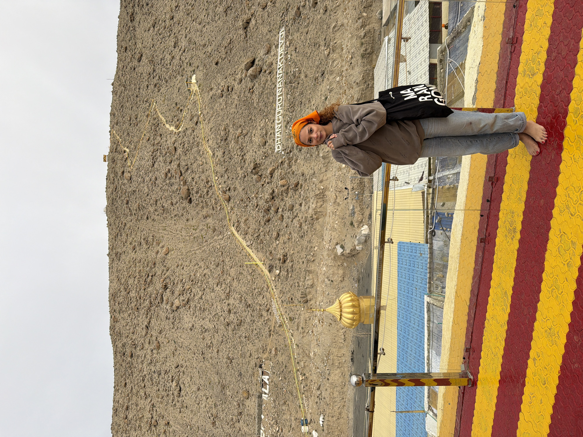 A young woman with curly brown hair and an orange headscarf stands on a raised platform in Chumkar, India. She is wearing a tan sweatshirt and light blue jeans, and is barefoot. She holds her hands clasped in front of her chest and carries a black tote bag with white lettering. The platform has a red and yellow striped pattern. Behind her, a steep, rocky hillside is visible, with a winding yellow path leading towards the top. Large white letters spelling "DHAN GURU NANAK DEV JI" are visible on the hillside. A golden dome structure is also present on the hillside, and a metal railing runs along the edge of the platform. The sky is overcast.
