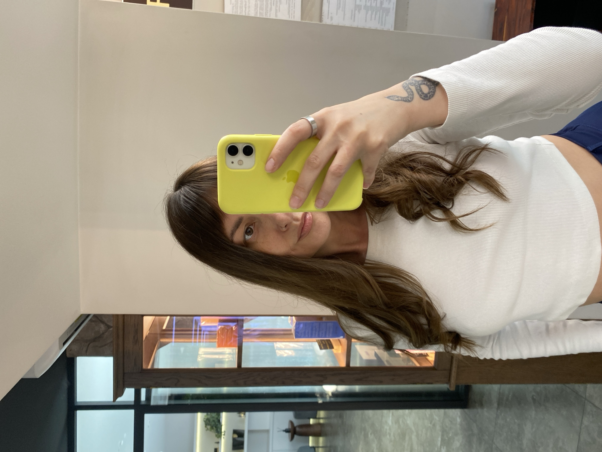 A woman with long brown hair and bangs, wearing a white ribbed long-sleeve top and dark blue bottoms, is taking a mirror selfie. She is holding a yellow iPhone in her right hand. A silver ring is visible on her right ring finger, and a snake tattoo is on her right forearm. She has a nose ring and freckles. The background shows a retail display cabinet filled with various products, with a large window and shelving units visible through it, suggesting a commercial or salon environment, potentially located in Votkinsk, Russia.