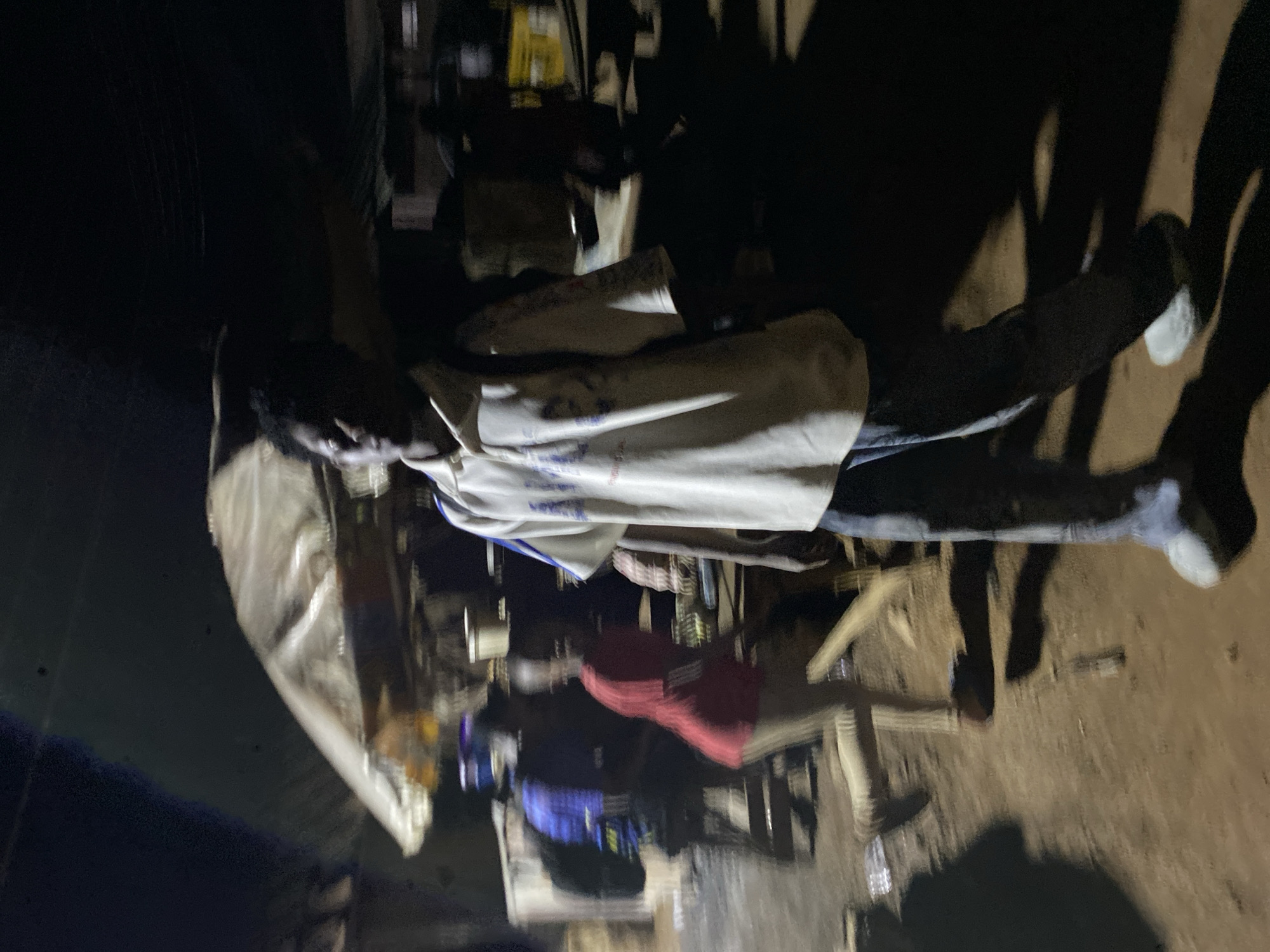 The image captures a nighttime scene, likely outdoors or in a semi-enclosed space, in Bukuru, Nigeria, illuminated by artificial light sources that cast long, distinct shadows on the ground. The low light conditions result in a somewhat grainy and blurry photograph.
The central subject is a man standing upright, facing slightly left of the camera. He wears a light-colored (possibly white or off-white) short-sleeved t-shirt with some blue and red, illegible text or a logo on the front. He is also wearing dark trousers, possibly jeans, and light-colored shoes. His hair appears dark and short. His right arm is slightly raised.
Several other individuals are present but less clearly visible. To the left and slightly behind the central man, another person wears a red shirt. Further back and to the left, a person in a blue shirt is partially seen. Other figures are obscured or only suggested by shadows.
The scene appears to be a casual gathering or a market-like setting at night. To the left of the central man, a large piece of white, possibly canvas or plastic, material is draped over an unseen structure, suggesting a stall or temporary shelter. In the background, various objects are clustered, including hints of yellow packaging. The ground beneath the individuals appears to be dirt or sand, and a plastic bottle can be seen lying on it in one of the crops. The strong artificial lighting creates dramatic shadows of people and structures on the ground. No discernible emotions are clear due to the image quality and distance.