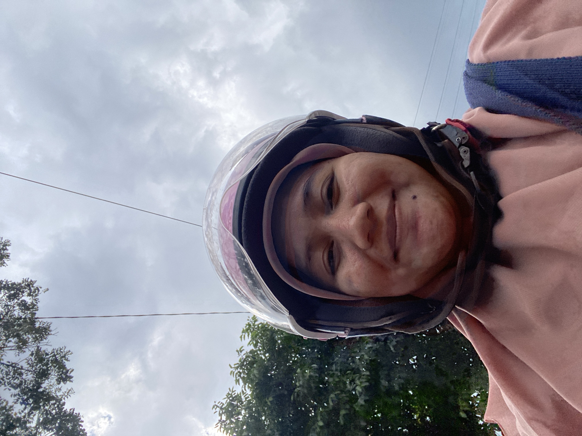 A young woman wearing a pink hijab and a black and pink helmet smiles into the camera. She is outside with trees visible to her left and a cloudy sky above. The scene appears to be in the city of Banjarbaru, Indonesia.