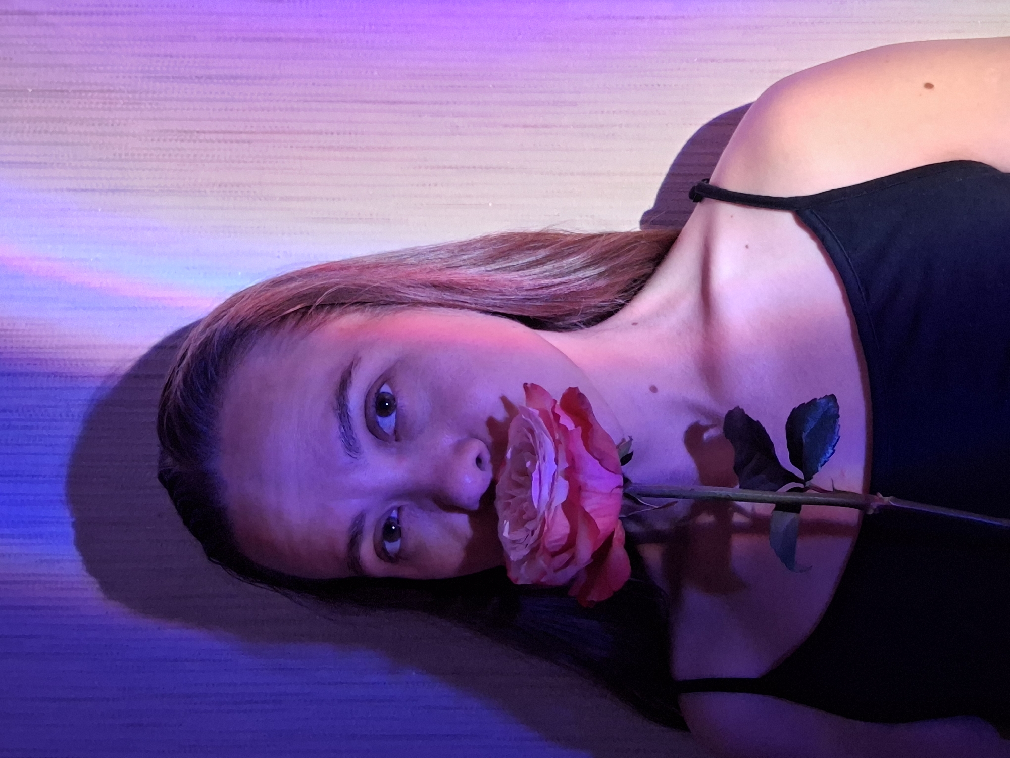 A woman lies horizontally, turned to face the camera, with a rose held in her mouth. She is wearing a black tank top. The lighting is a mixture of pink, purple, and blue, creating a dramatic and slightly surreal effect. The background appears to be a wall or surface with subtle horizontal lines. No specific details in the image point to the city of Chaikovsky, Russia.