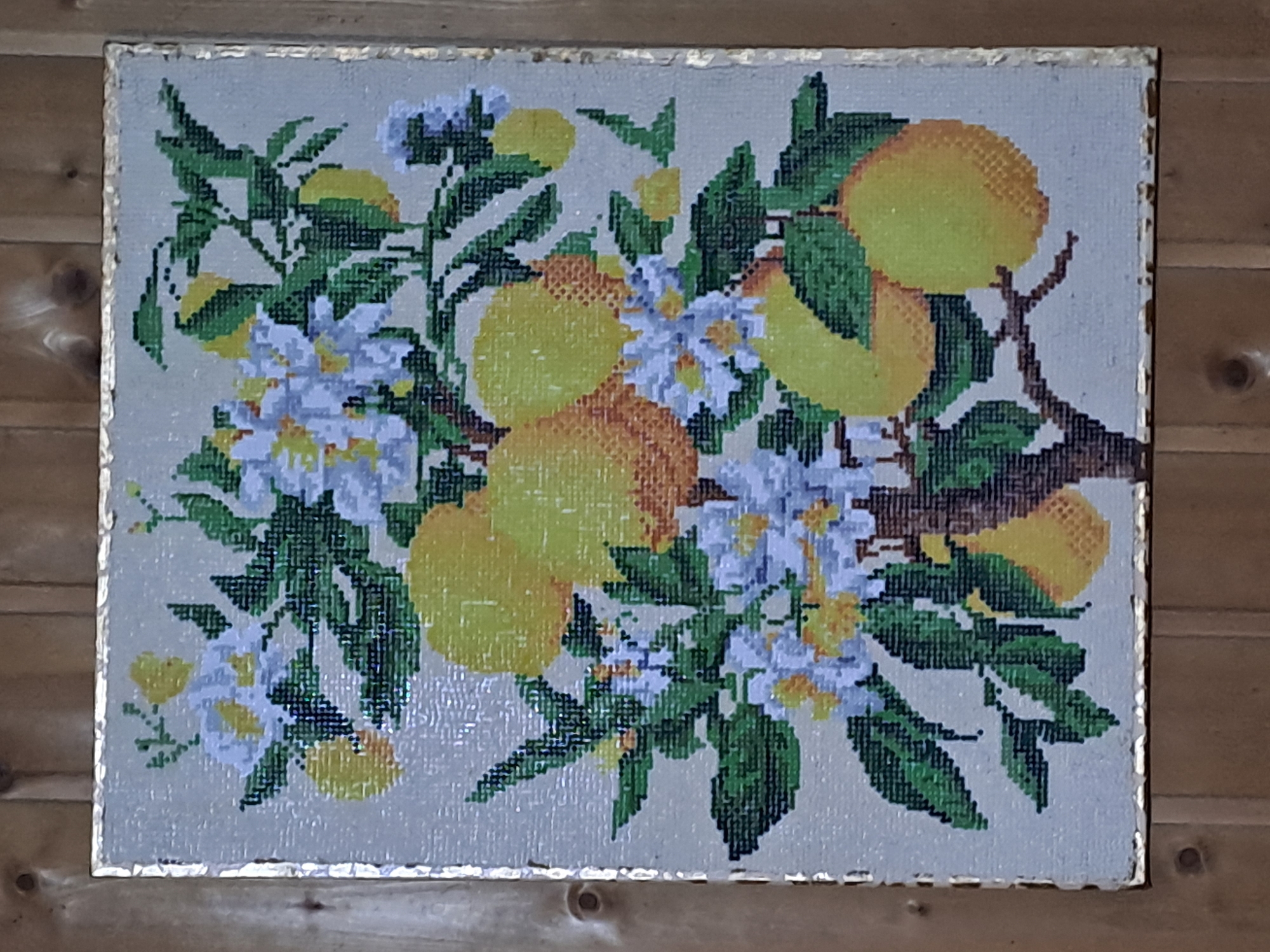 A framed cross-stitch depicts a lemon tree with vibrant yellow lemons and delicate white and yellow blossoms against a pale background. The artwork is positioned on a wooden surface, likely a wall, characterized by vertical planks and visible knots. The frame of the artwork has a subtle gold sheen.
