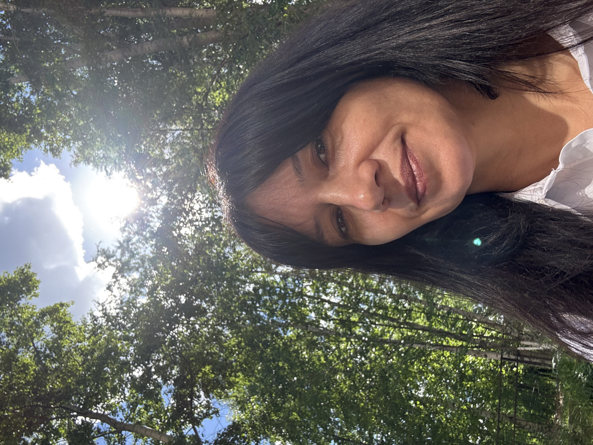 A selfie taken from a low angle shows a woman's face and upper body against a backdrop of trees and sky. The woman has long, dark hair and is smiling. The sun is shining brightly through the leaves of the trees, creating a lens flare effect. The environment appears to be a park or wooded area. The image does not contain any information that would allow for its location in the city of Chaikovsky, Russia.