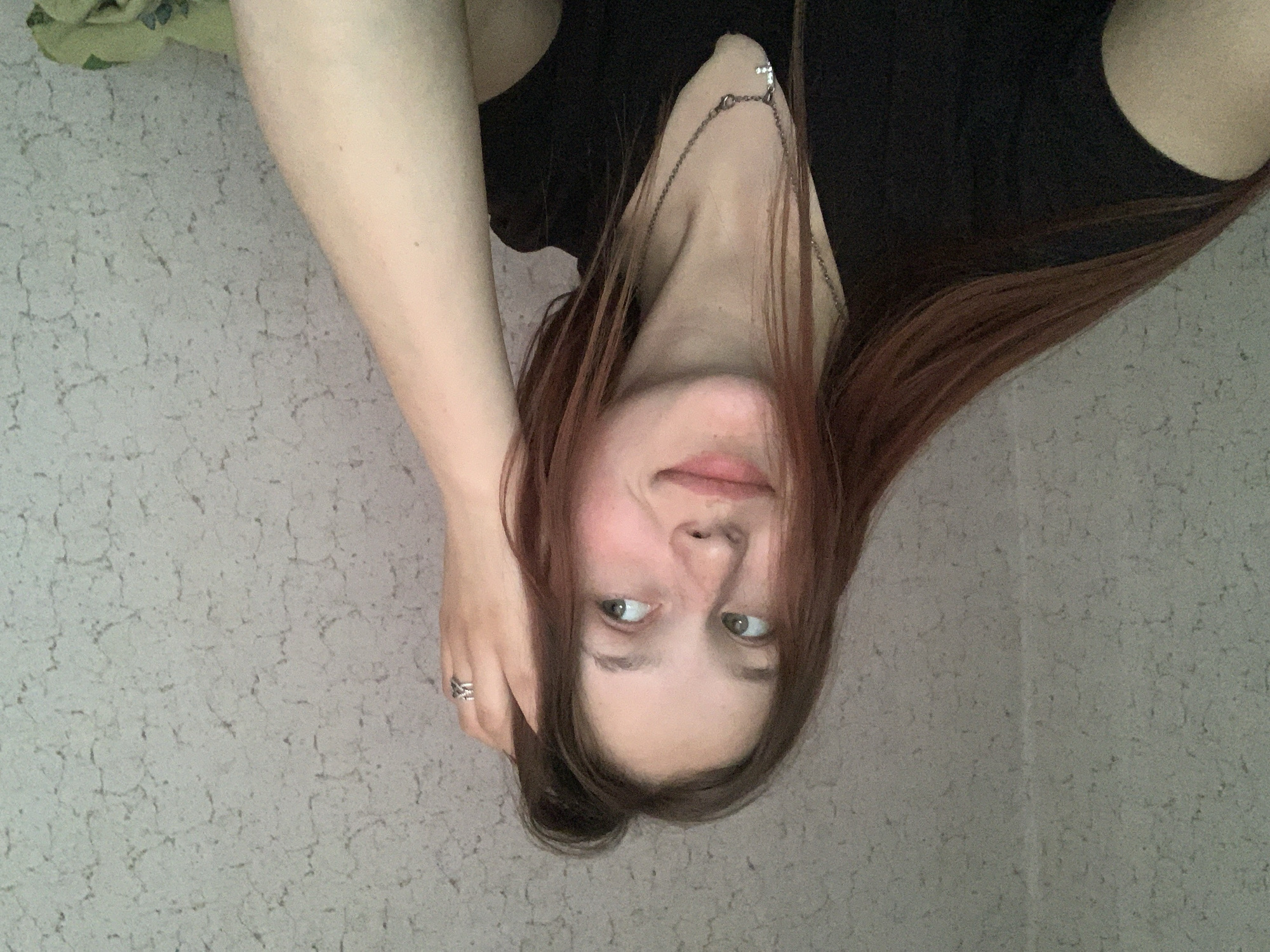 A young woman is photographed from an upside-down perspective. She has fair skin, light green eyes, and reddish-brown hair. She is wearing a black top and a silver necklace with a pendant. Her left hand, adorned with a silver ring, is near her head. The background features a textured, light-colored wall and a glimpse of a green object, possibly a pillow or piece of furniture, at the top of the frame. There is no indication of Фоки, Russia.