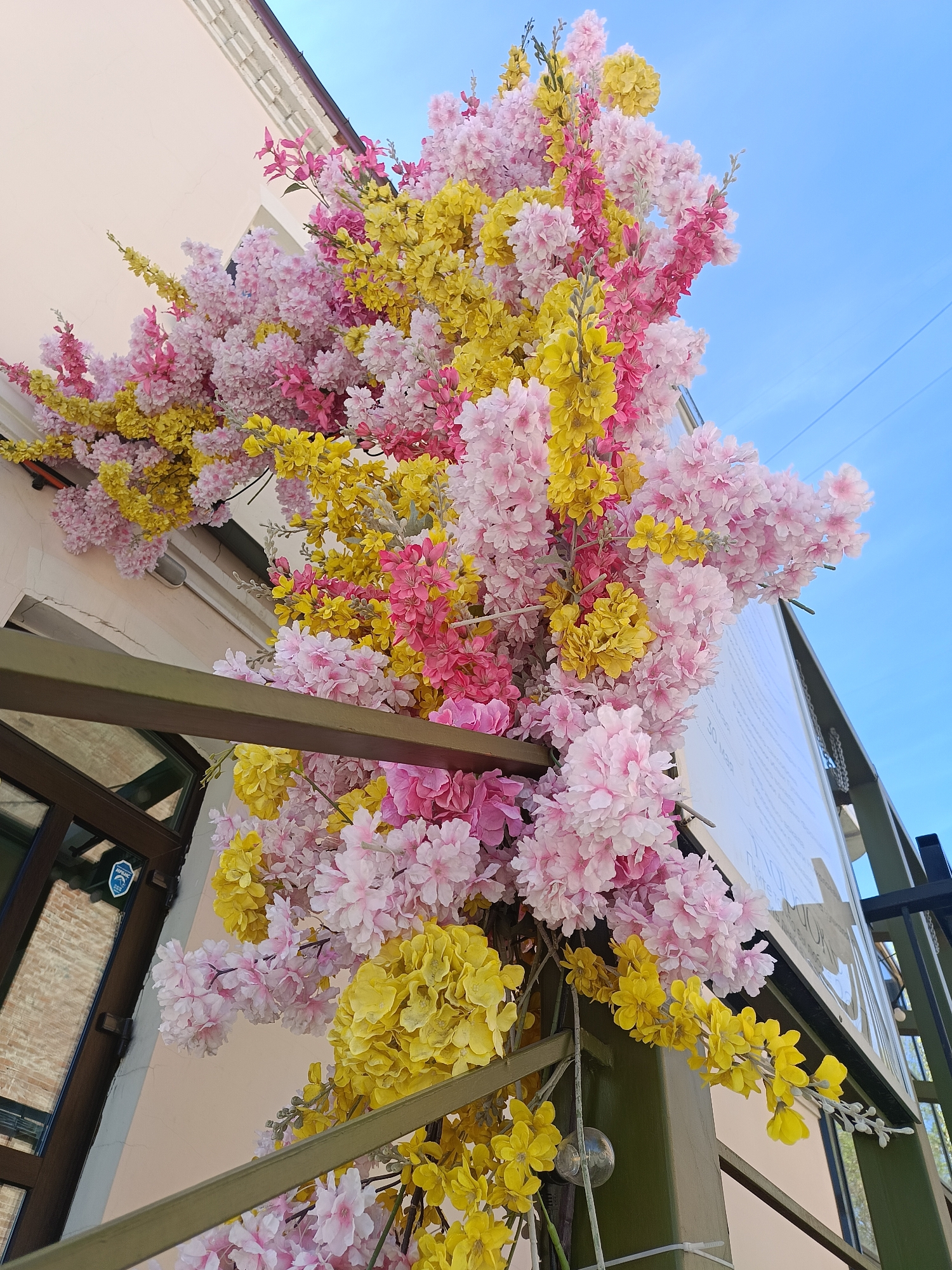 A low-angle shot shows a building facade decorated with pink and yellow artificial flowers. The flowers are attached to a metal structure near a building entrance with a glass door bearing a security sticker. The sky above is blue. This scene is located in Izhevsk, Russia.