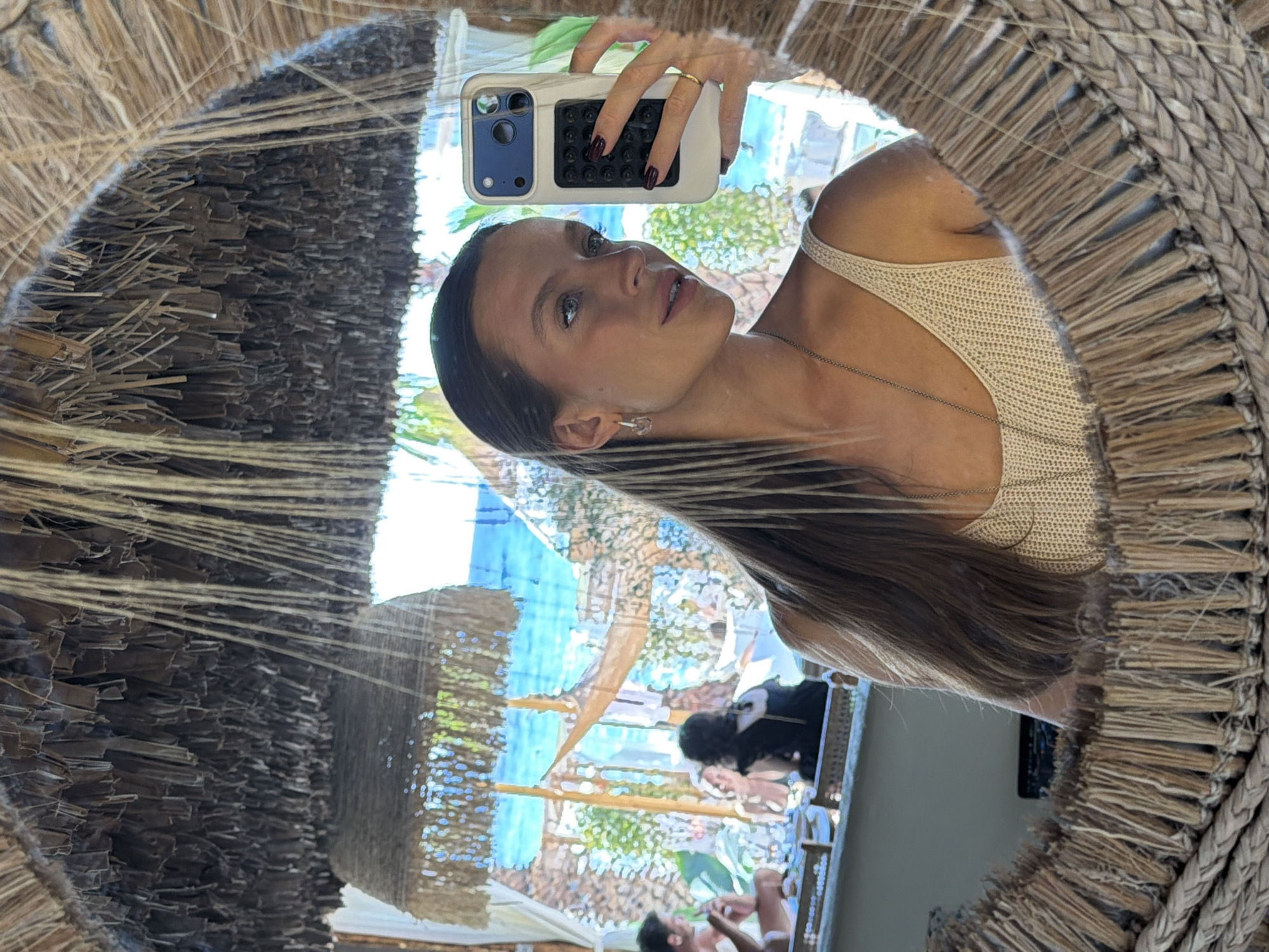 A woman is taking a selfie in a mirror at a restaurant in Drobnići, Montenegro. She is wearing a beige knit tank top and a silver necklace. She has long, dark brown hair and is wearing earrings and a ring. The mirror is framed with natural materials, likely rattan or straw. In the background, other people are visible sitting at tables, and beyond them is a body of water, likely the Adriatic Sea, and palm trees, suggesting a coastal location. The atmosphere appears relaxed and summery.
