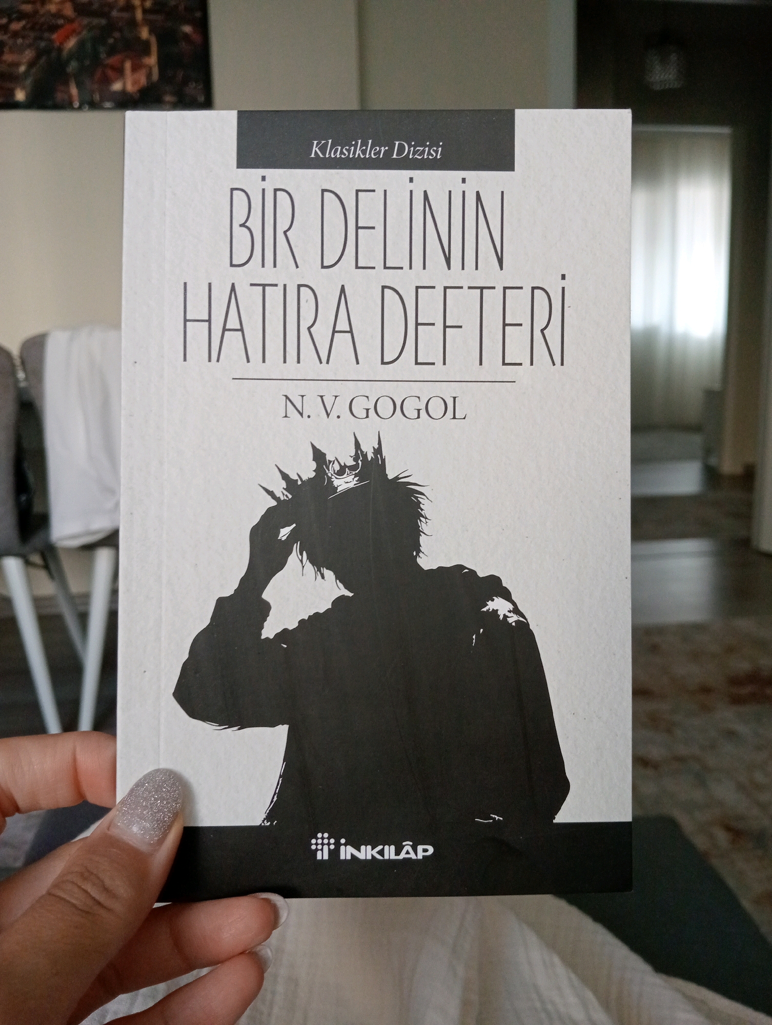 The image shows a close-up of a book cover titled "BİR DELİNİN HATIRA DEFTERİ" (Diary of a Madman) by N.V. GOGOL. The cover features a silhouette of a person wearing a crown and holding their head. The book is white with black text and graphics. A person's hand with glittery nail polish is visible in the foreground, holding the bottom left corner of the book. The background appears to be an indoor setting, possibly a living room, with furniture and a window with sheer curtains visible. There is no indication or information within the image that suggests any connection to the city of Silifke, Turkey.