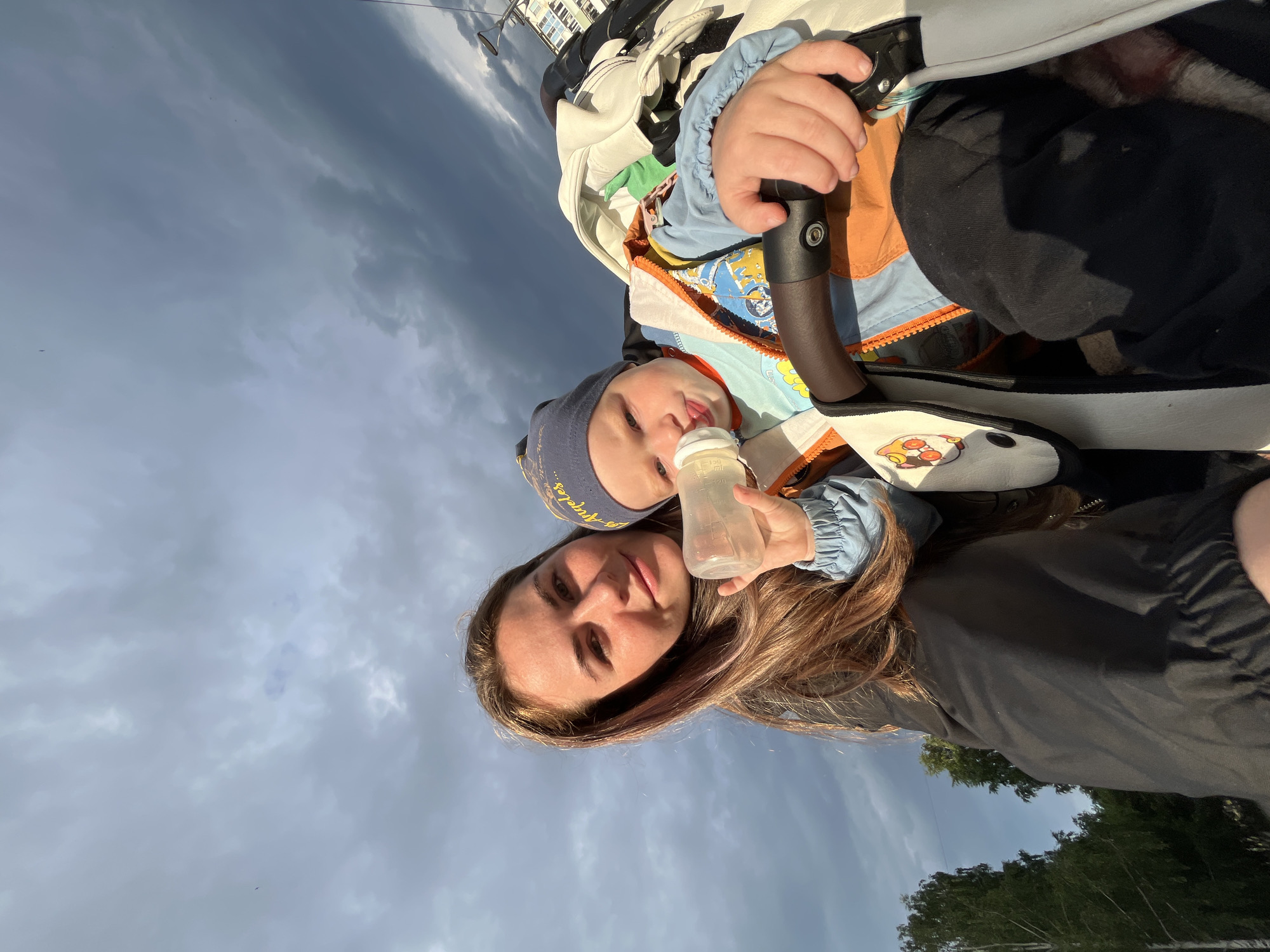 The media file features a selfie of a woman and a baby outdoors. The woman, with long, reddish-brown hair, is in the foreground, looking towards the camera with a gentle expression. She is wearing a black jacket. Beside her, a baby is seated in a stroller, wearing a blue cap and a colorful jacket. The baby is holding a clear baby bottle with a white nipple and appears to be drinking from it.
The background is dominated by a cloudy sky, suggesting overcast or stormy weather. Some trees and a building are visible at the bottom of the frame, indicating an outdoor setting. The overall lighting suggests it might be late afternoon or early evening, as the sun is low.
The image appears to be taken in Chaikovsky, Russia. No visible text is present in the image itself, other than markings on the baby bottle which are not legible. The mood conveyed is warm and intimate, capturing a moment between a mother and child.