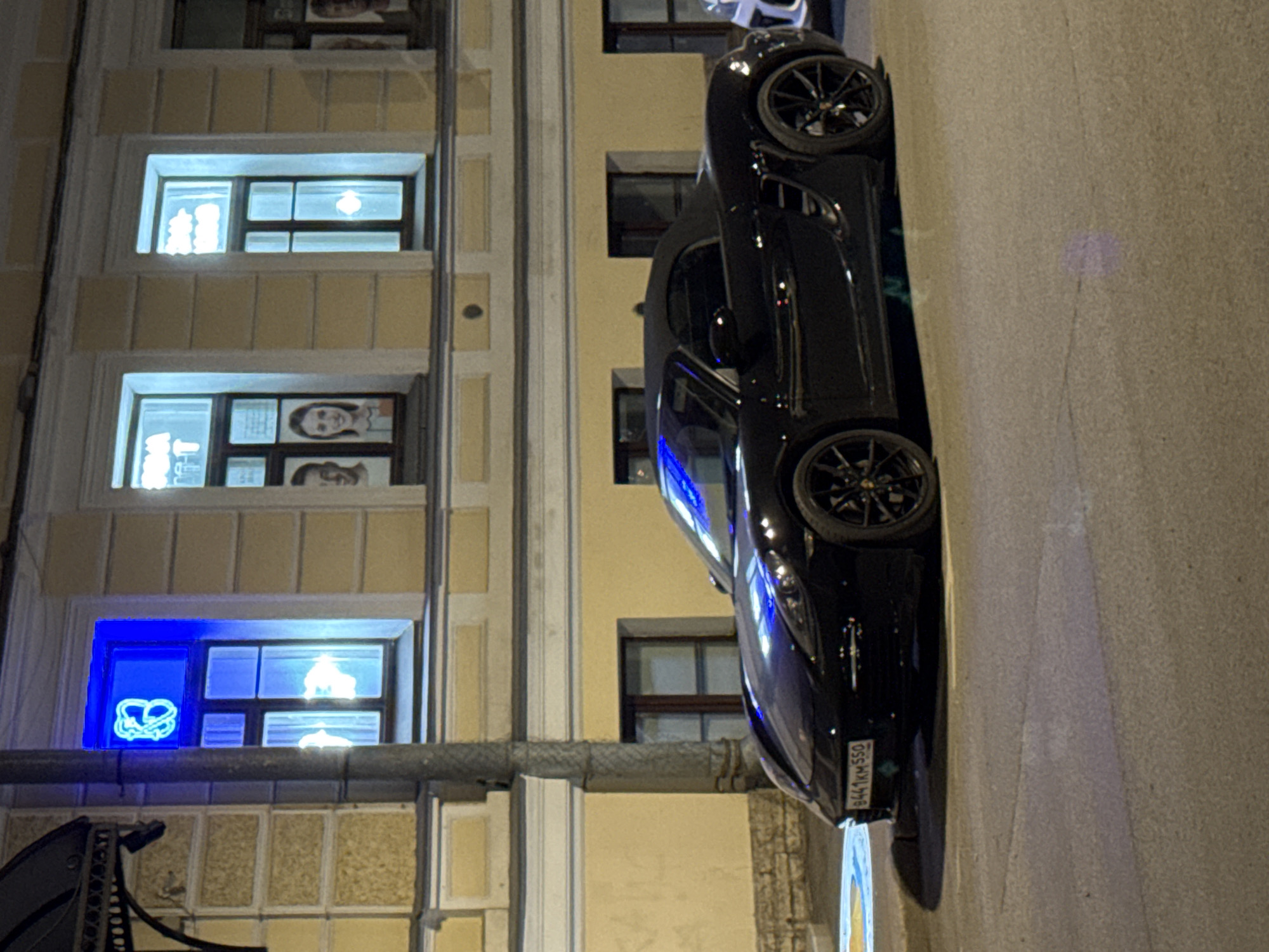 The image depicts a nighttime scene in Saint Petersburg, Russia, focusing on a black convertible car parked alongside a building. The car's license plate is visible, reading "B441KM550." The building has several windows, some illuminated. One window has a blue light with a pretzel-shaped neon sign. Other windows display images of people, appearing to be advertisements or signage. The street is dimly lit, with a visible sidewalk or road surface. A portion of a street lamp or pole is visible in the lower left. The scene is typical of an urban environment at night, with the architectural details characteristic of Saint Petersburg.