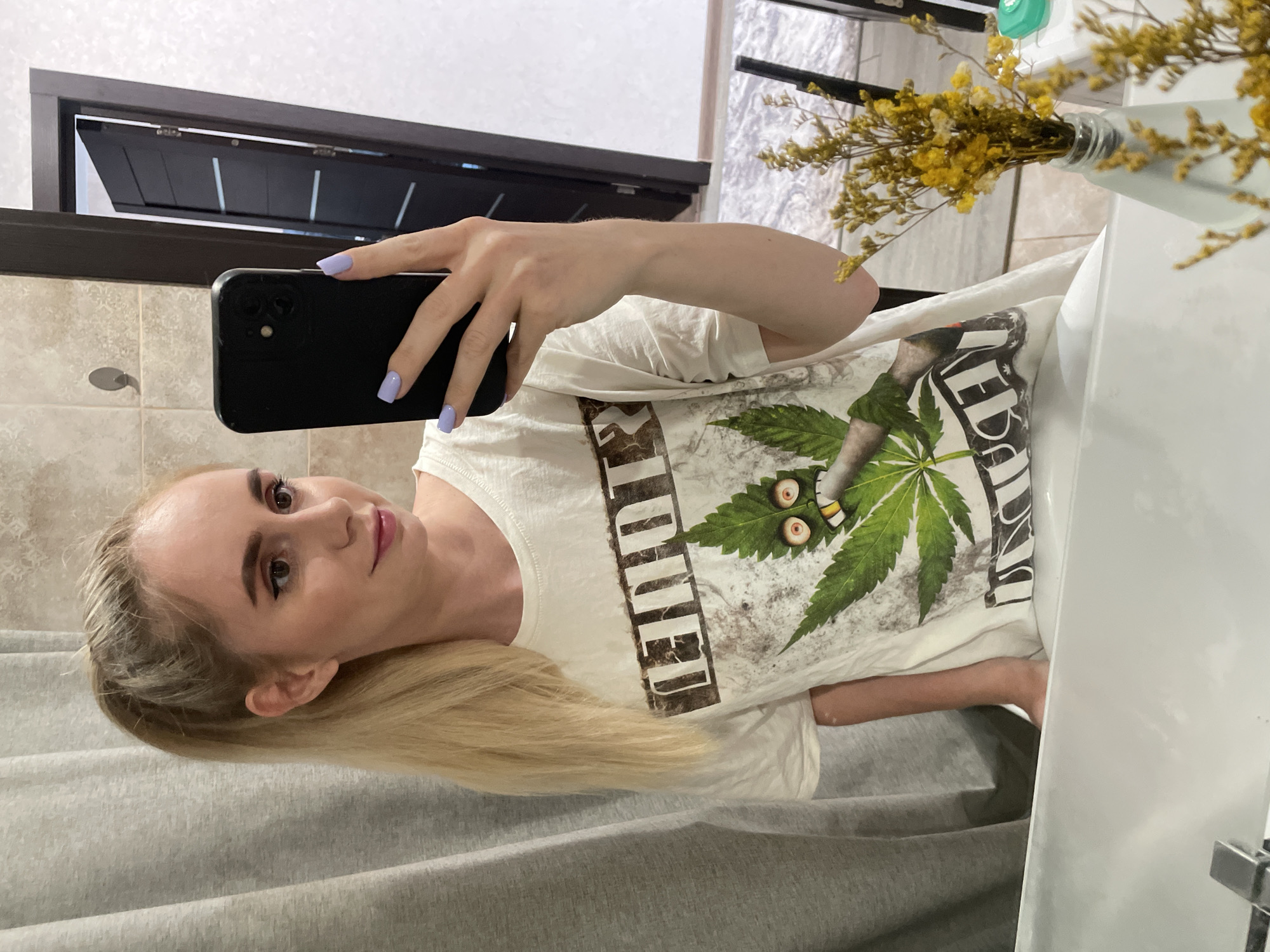 A young woman is captured in a mirror selfie, holding a dark smartphone in her right hand, which displays light lavender-painted fingernails. She has long, straight blonde hair and is wearing makeup, including eyeliner and light pink lipstick, with a neutral to slightly smiling expression as she looks towards her phone. She is dressed in a light-colored, likely white, short-sleeved t-shirt.
The t-shirt features a graphic design with a prominent green marijuana leaf adorned with cartoonish, bloodshot eyes and a mouth, alongside a lit joint or cigarette. Partial text on the shirt reads "LEGALIZE" vertically on the left and "WEED" on the right.
The setting appears to be a bathroom, characterized by a white sink basin visible on the right and light beige square-tiled walls in the background. A light gray shower curtain or bath mat is present in the foreground. In the reflection of the mirror, a dark wooden door frame with a partially open door is visible, suggesting an adjoining room. A frosted glass vase containing dried yellow flowers rests on the counter near the sink. The image is oriented sideways, implying a vertical original capture. The indoor scene is illuminated by artificial light. The location is Vladivostok, Russia.