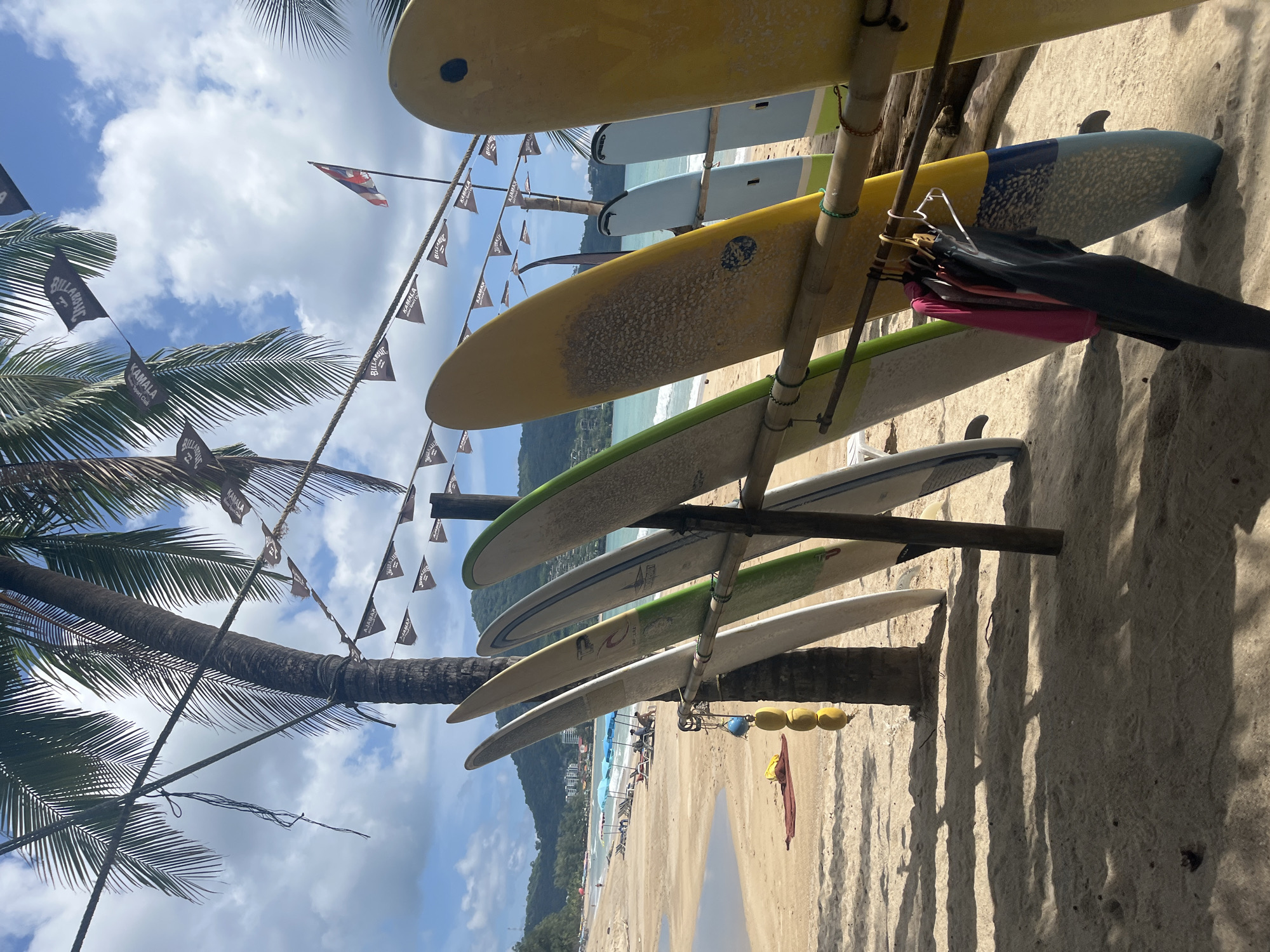 In Ban Bang Thao, Thailand, a row of surfboards stands upright on a sandy beach, leaning against a bamboo rack. The surfboards vary in color, with some displaying yellow, green, and white hues. Clothing items, including a pink shirt and dark pants, hang on hangers attached to the rack below the surfboards. In the background, a tropical beach scene unfolds with palm trees, beach umbrellas, lounge chairs, and the ocean under a partly cloudy sky. A small flag is visible on a pole in the distance. The ground is covered in sand, with shadows cast by palm fronds.