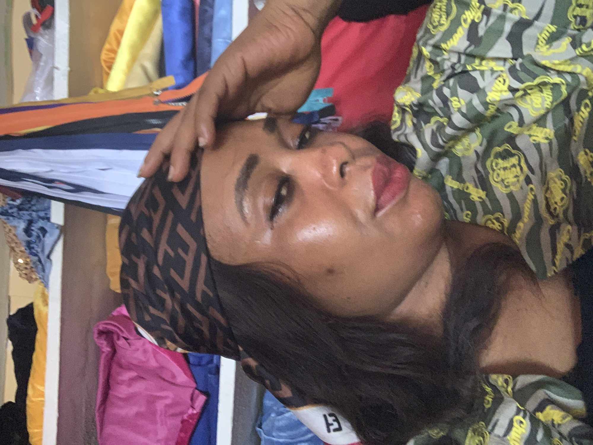 An indoor portrait features an adult female, positioned with her head tilted, looking directly at the camera. She has a medium-dark skin tone, dark hair, and her right hand rests gently on her forehead. She is wearing a dark brown and black patterned headwrap, which appears to have a repeated 'F' logo, possibly indicative of the Fendi brand. Her top is a camouflage-style garment with green, grey, and black patterns, featuring numerous yellow logos that clearly read "Chupa Chups." A black garment is visible beneath her camouflage top.
The background is filled with various colorful fabrics and tailoring supplies, suggesting a textile shop, market stall, or workshop. Rolls or piles of materials in hues of yellow, blue, pink, red, black, and purple are visible. Long, colorful zippers in orange, purple, and black are also present, some appearing to be stacked or displayed. The lighting suggests it is daytime. The overall scene is static, with the subject posing for the photograph. The setting is consistent with a commercial establishment in Sabon Tasha, Nigeria.
