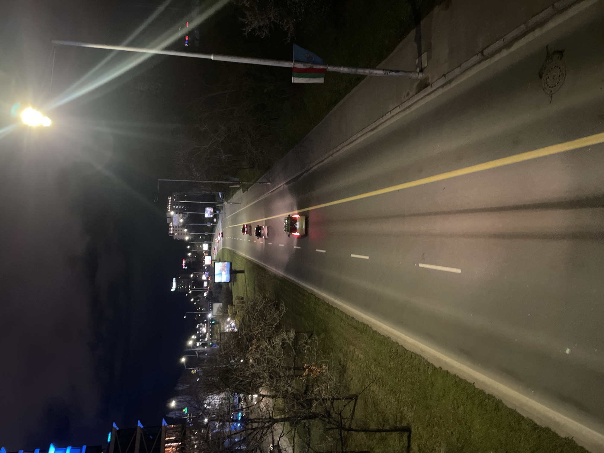 The image shows a night view of a multi-lane road in Sofia, Bulgaria, illuminated by streetlights and car headlights. Several cars are driving on the road. The road is marked with yellow and white lines. On the left side of the road, there is a sidewalk and a grassy area with trees. In the background, there are buildings with lights and billboards. A flag of Adygea is attached to a pole alongside the road. The sky is dark and cloudy. A manhole cover is visible on the right side of the road.